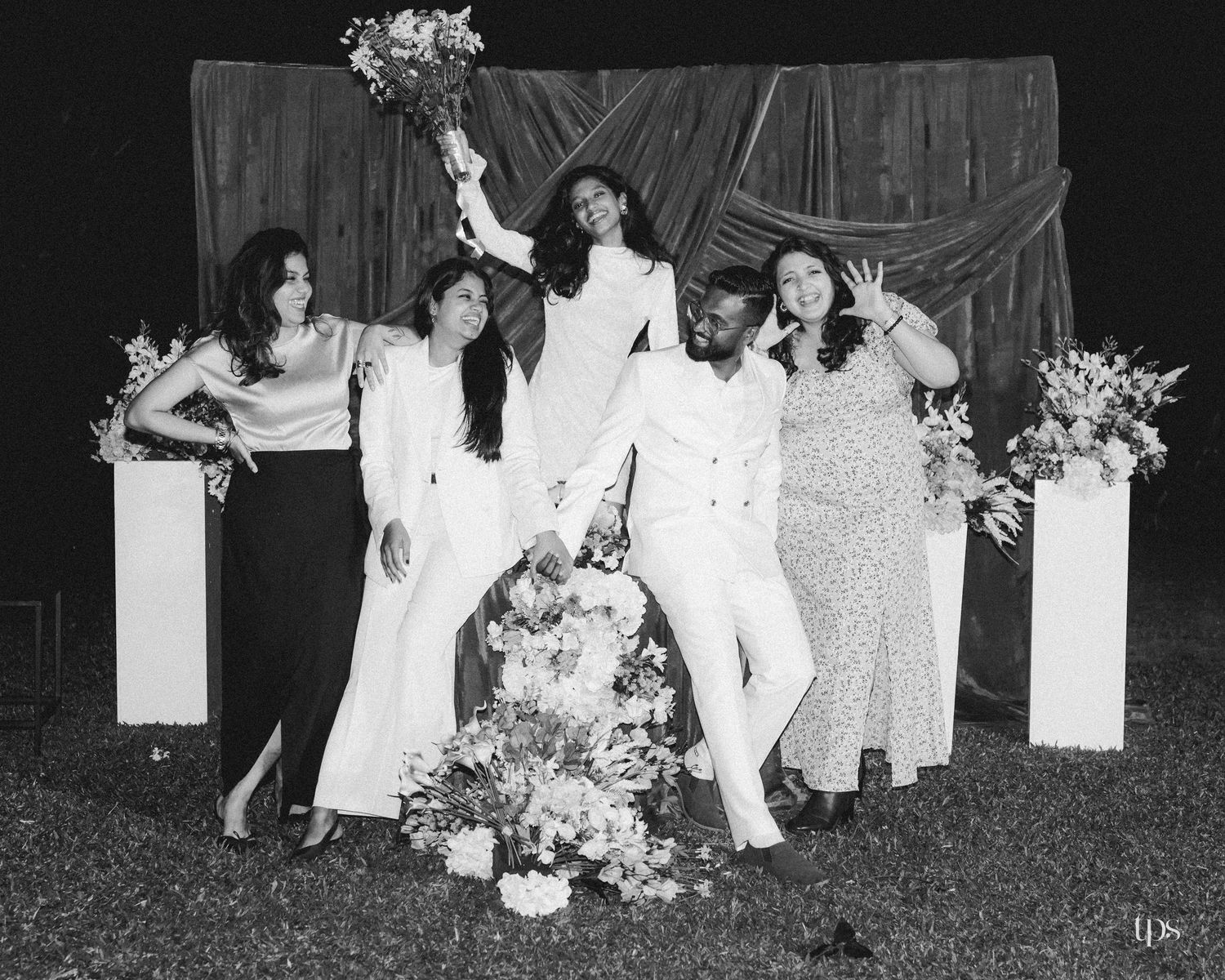 Black and white wedding photo of five joyful people posing with flower arrangements and a raised bouquet before a draped backdrop outdoors.