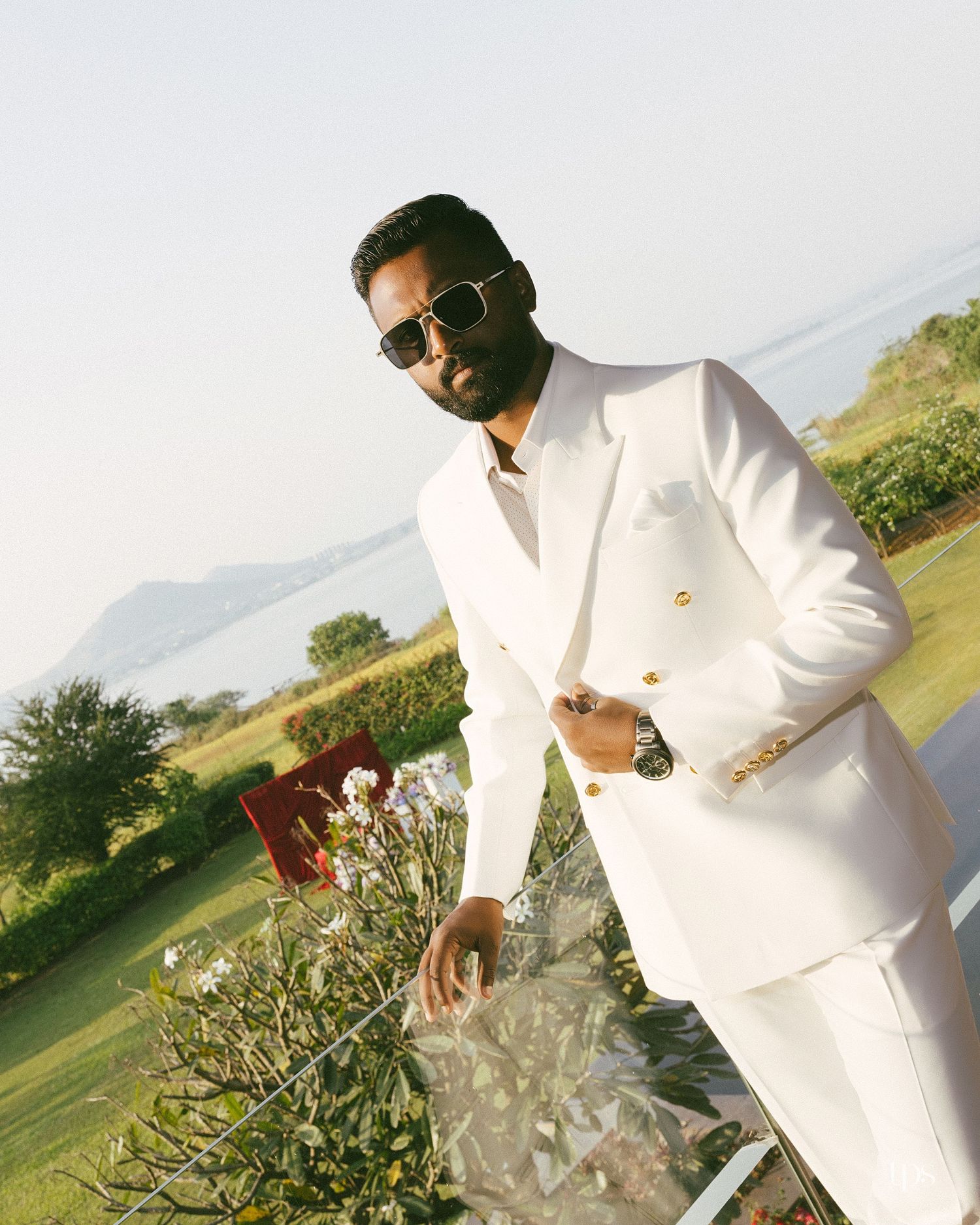 Man in sunglasses and a white double-breasted suit with gold buttons stands near flowers on a terrace overlooking a lake and hills.