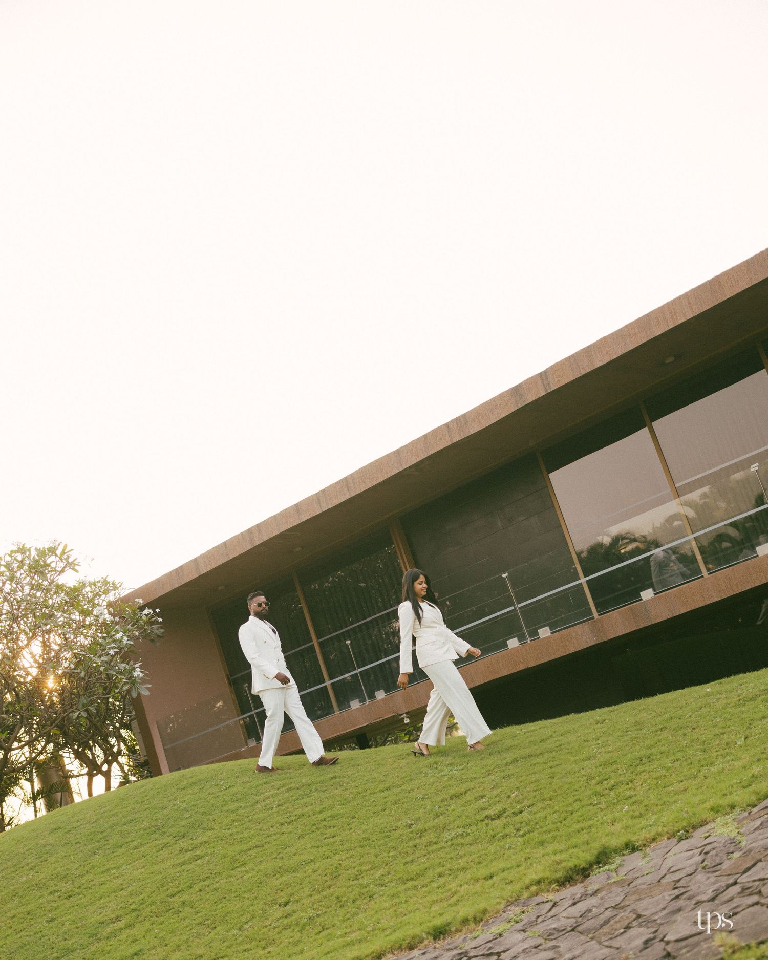 Two people in white outfits walk across a sunlit grassy hill beside a modern building with large glass windows and a sloped roof.