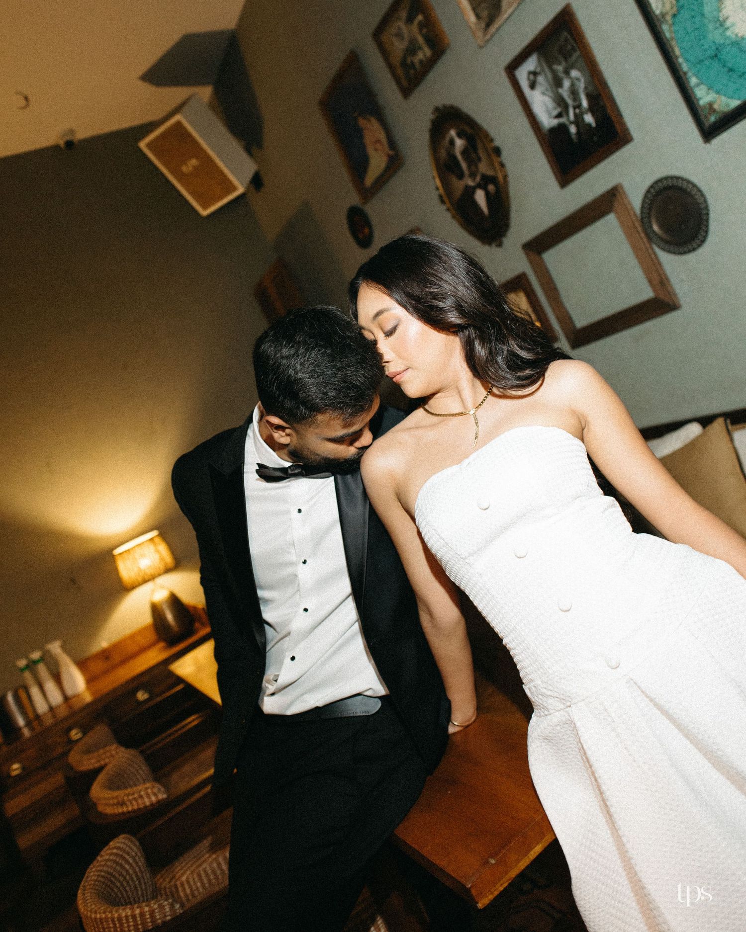 Groom in a black tuxedo and bow tie leans affectionately toward a bride in a white strapless dress in a cozy room with framed wall art.