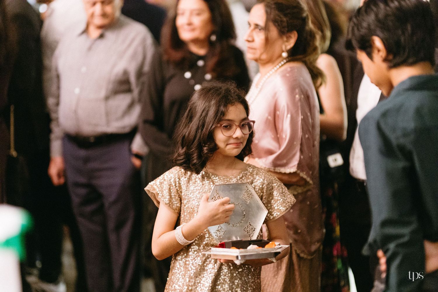 Young girl in glasses and a sparkly gold dress holds a plate of food while smiling guests gather at an indoor celebration behind her.