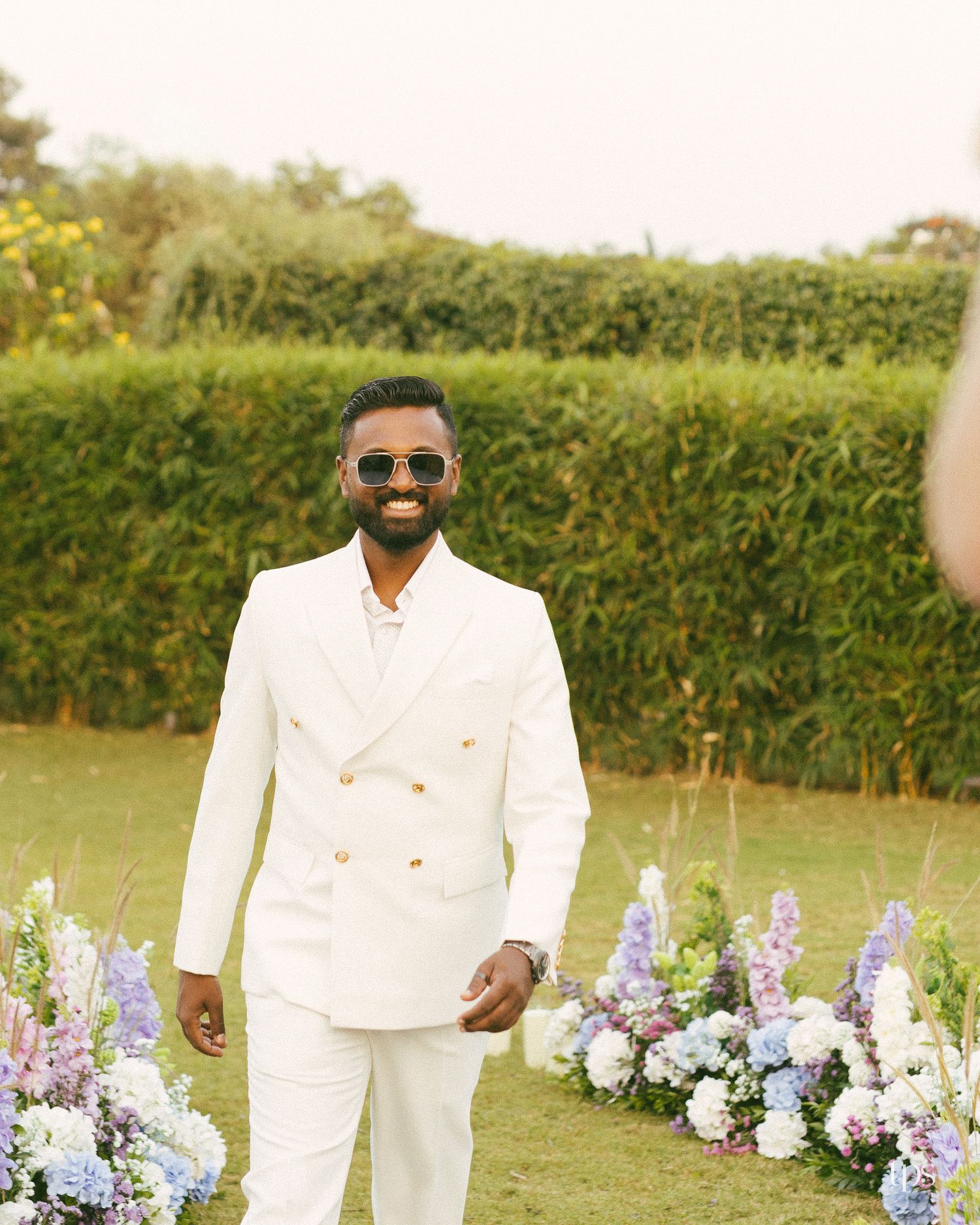 Smiling bearded man in sunglasses and a white double-breasted suit walks past blue and white flower arrangements on a sunny outdoor lawn.