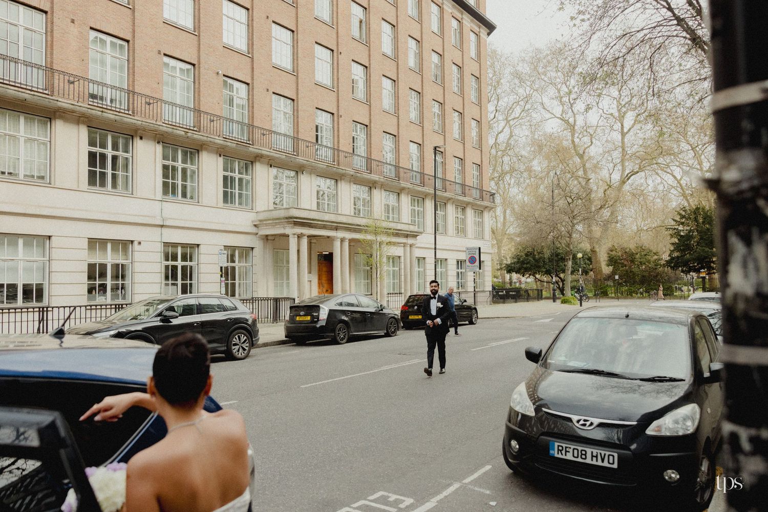 A wedding couple arrives outside a grand London building, with a shirtless groomsman and suited guest on a quiet street.