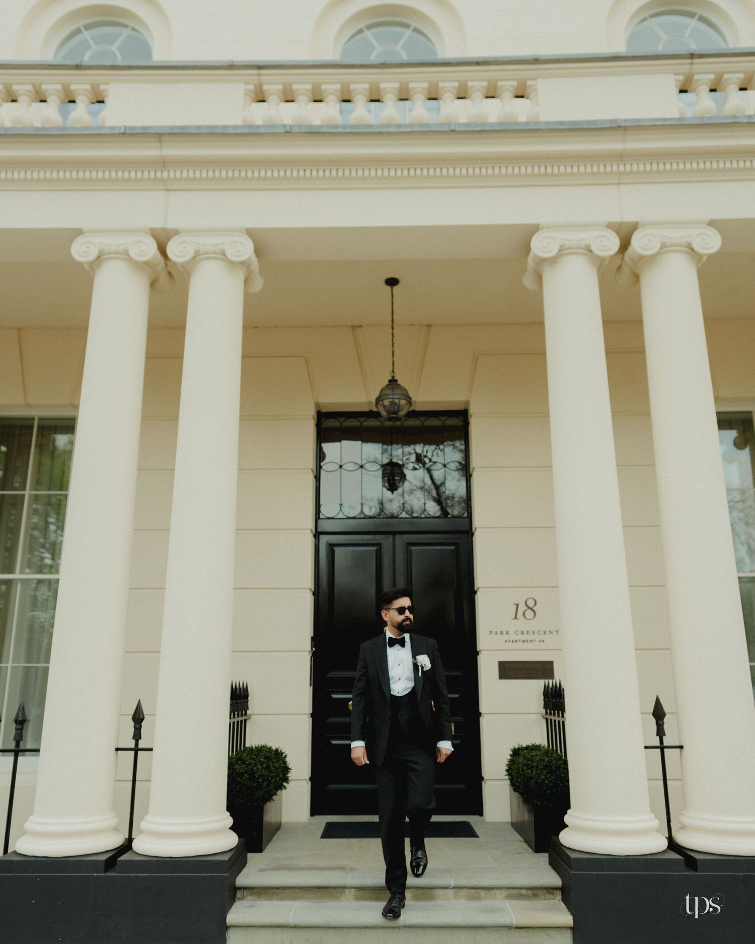 Groom in black tuxedo and bow tie stands on steps of elegant white neoclassical building with columns.