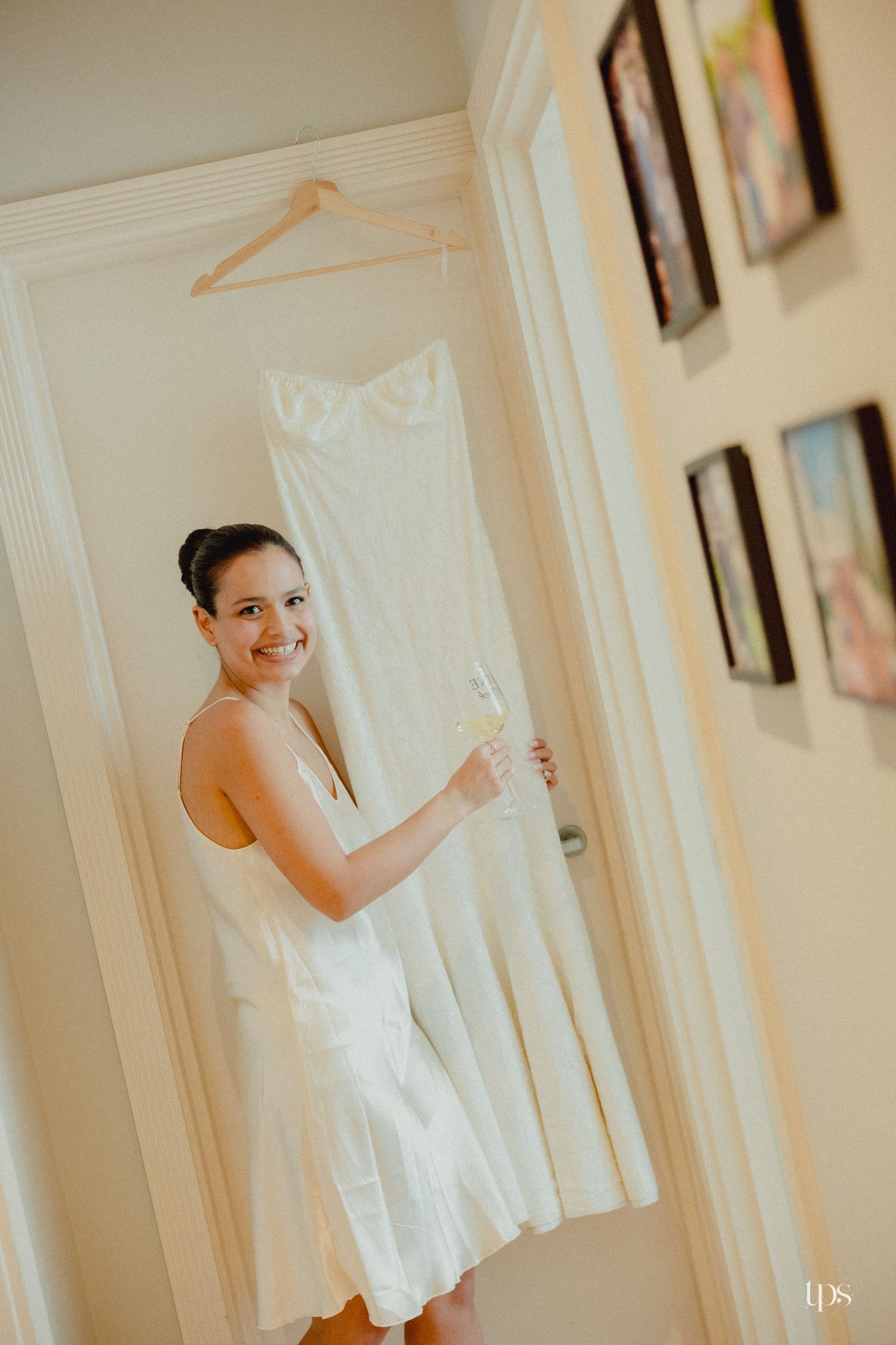 Smiling bride in white slip dress holding champagne glass standing in doorway with wedding gown hanging behind her.