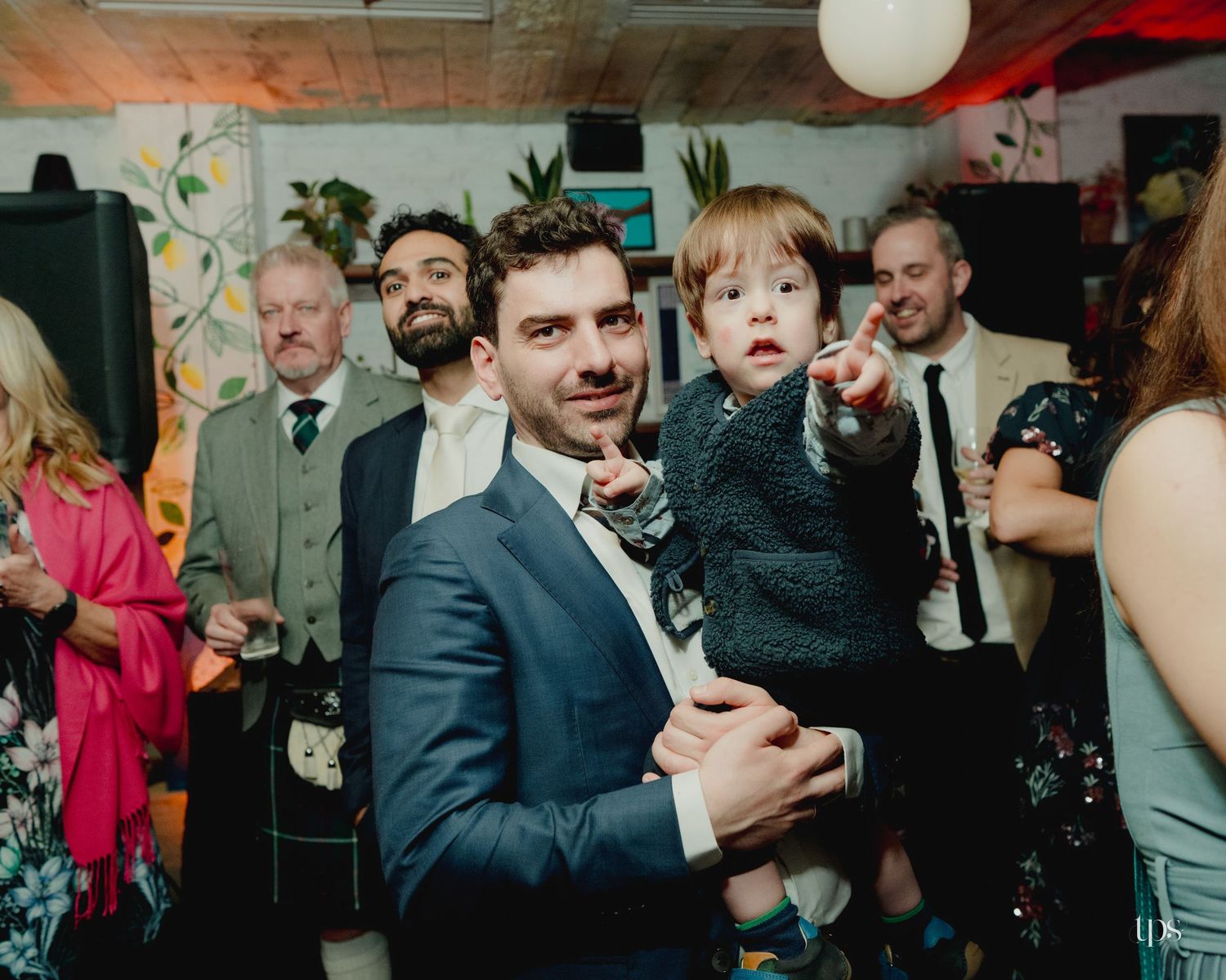 Man in blue suit holding a young child at a wedding reception party with guests in background.