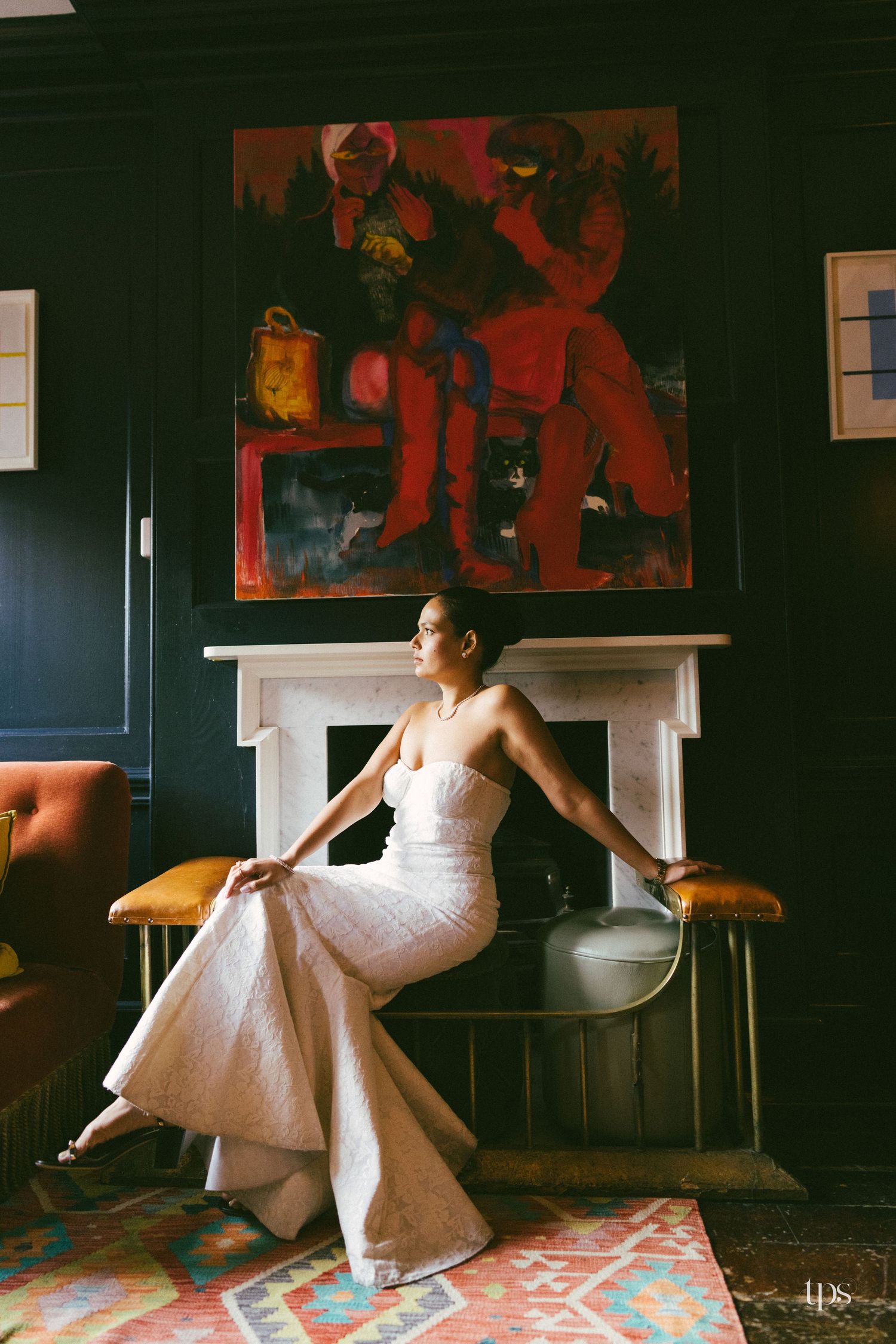 Bride in white strapless gown seated by fireplace with vibrant abstract painting backdrop in dark elegant room.
