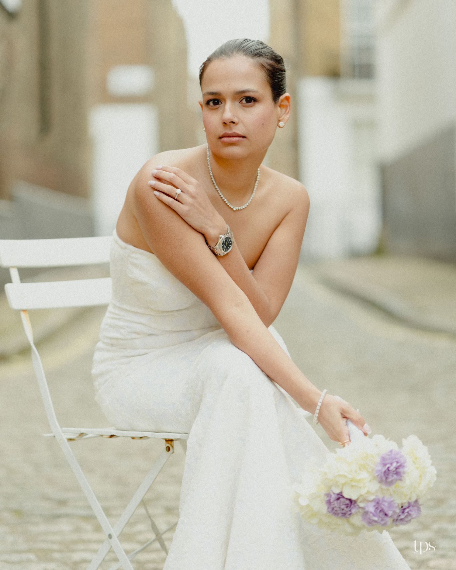 Elegant bride in white strapless gown sits on chair holding purple bouquet, wearing pearl necklace and watch.