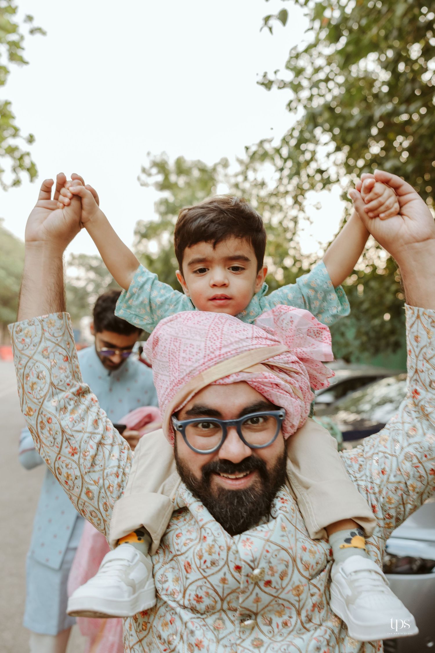 A bearded man in traditional Indian attire and pink turban carries a young child on his shoulders outdoors.