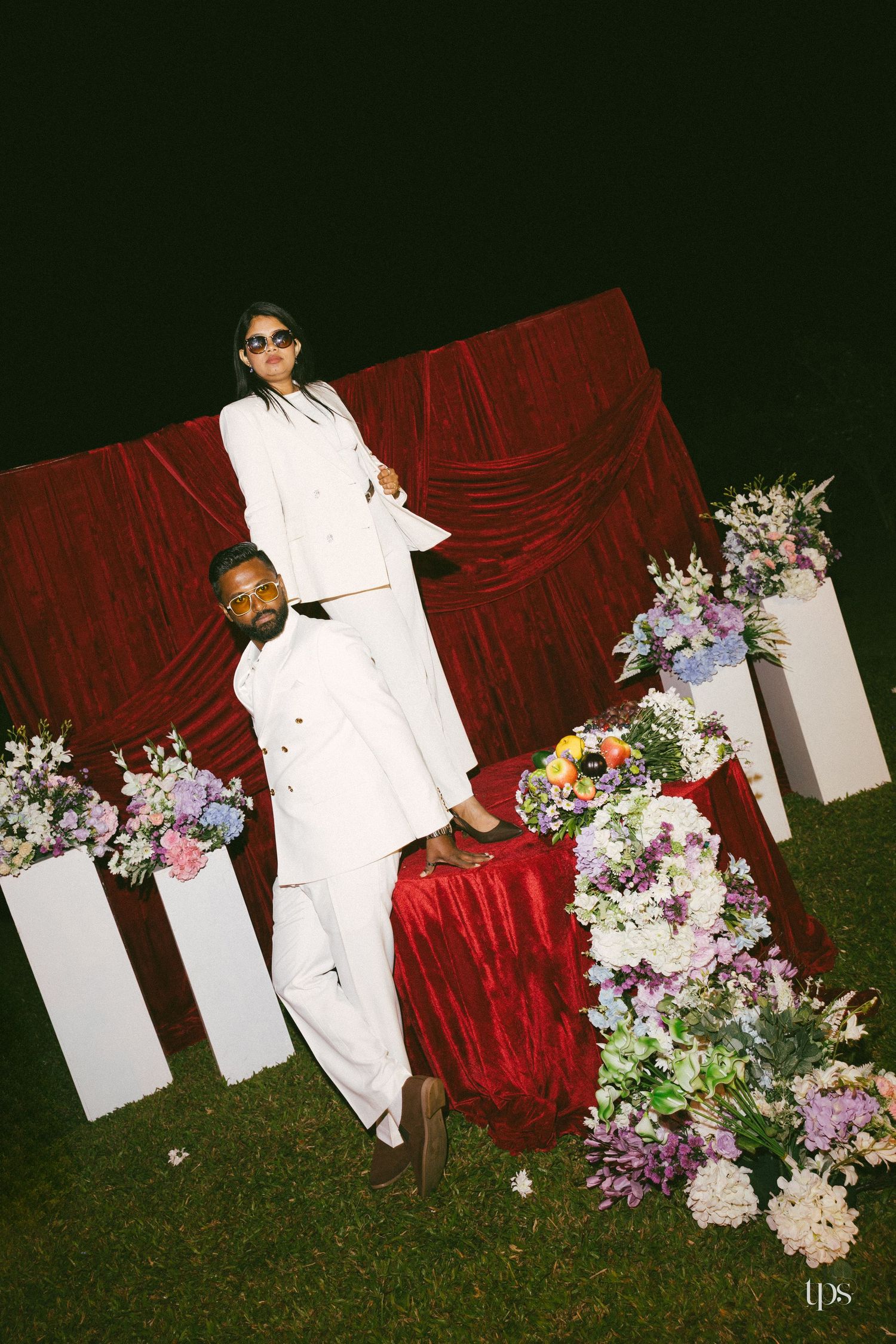 Two people in white suits and sunglasses pose outdoors at night beside a red velvet backdrop and pedestals of purple and white flowers.