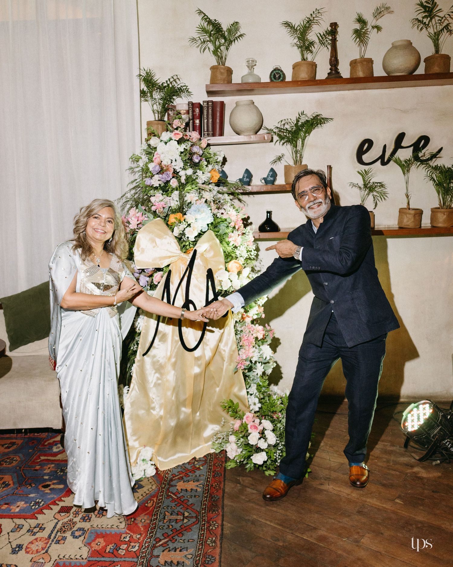 A smiling couple in elegant attire pose beside a floral arrangement with a monogram ribbon in a cozy room with shelves of potted plants.