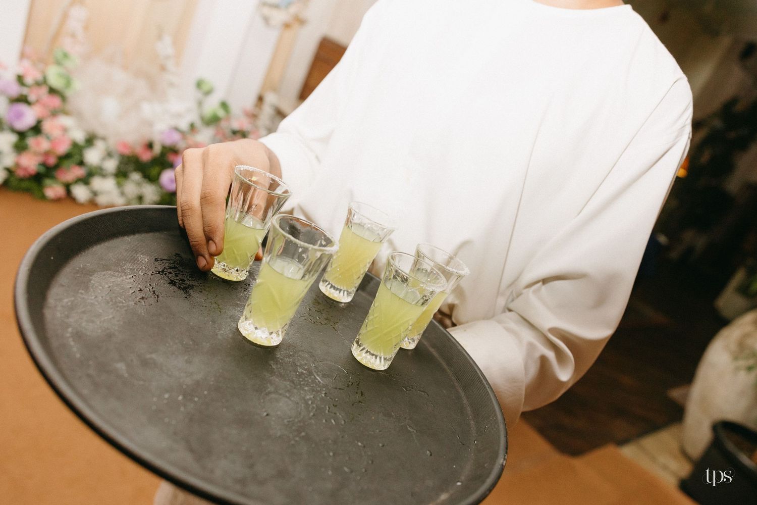 Server in a white outfit holds a dark round tray of small glasses filled with pale green drinks beside a floral arrangement at an event.