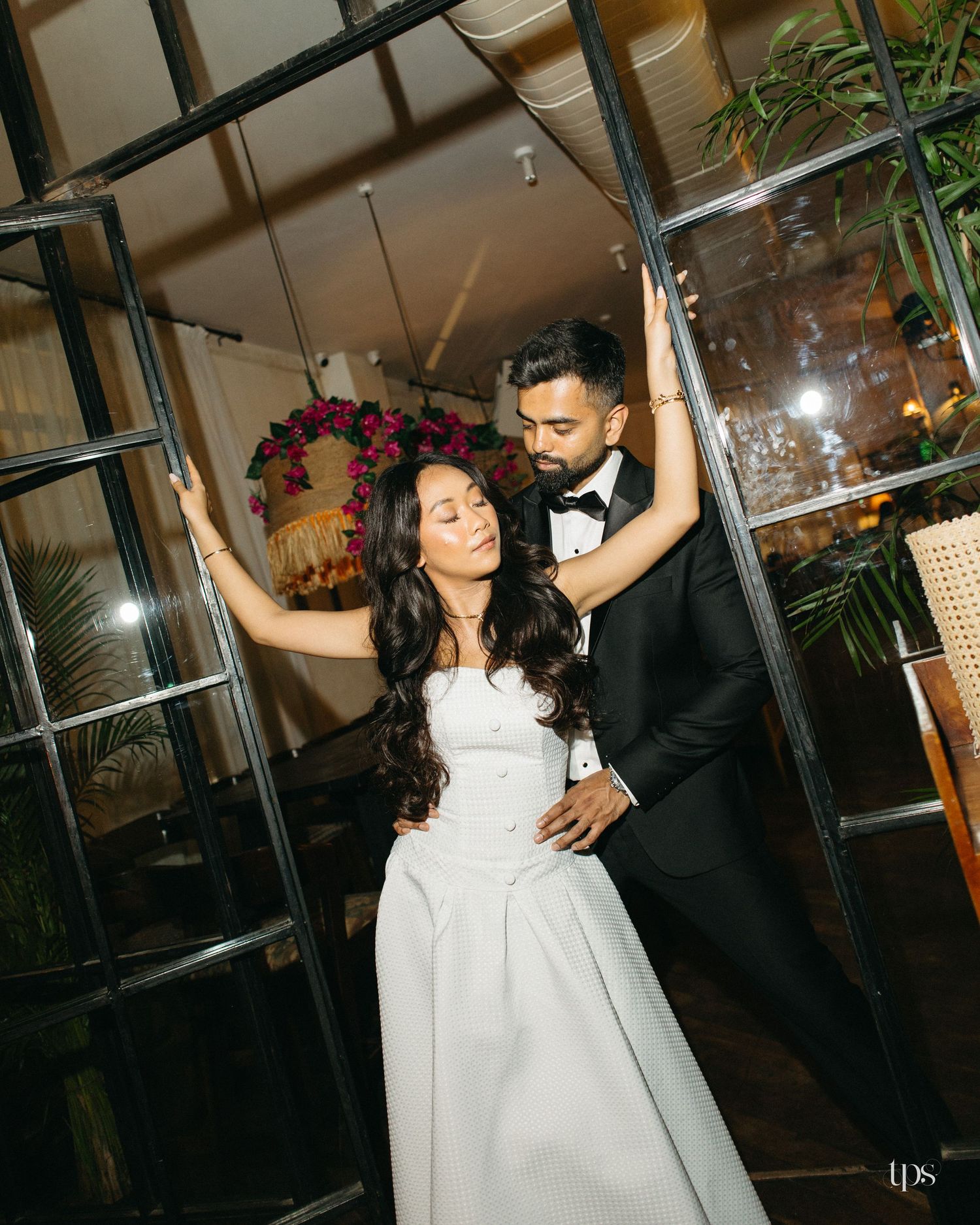 Bride in a white strapless gown and groom in a black tuxedo pose together between black-framed glass doors at an elegant wedding venue.