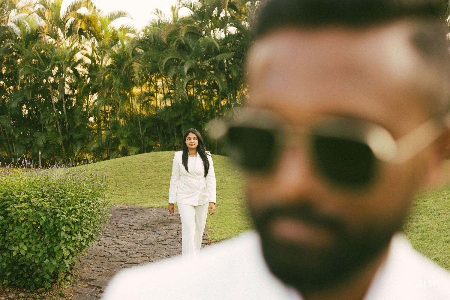 Blurred bearded man in sunglasses and white shirt in the foreground with a woman in a white suit standing on a garden path behind him.