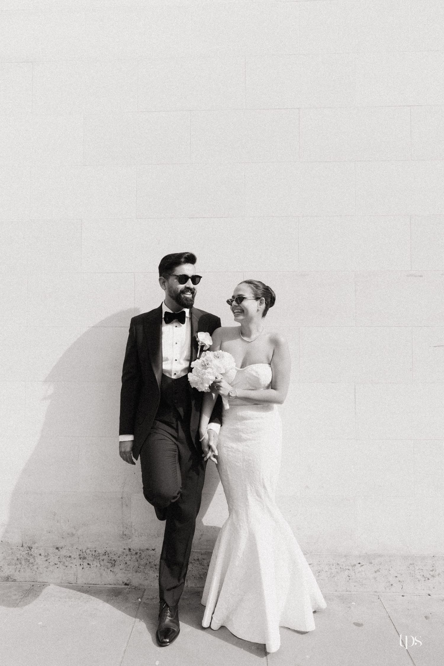 Black and white photo of a bride and groom in sunglasses leaning against a white brick wall, holding a bouquet.