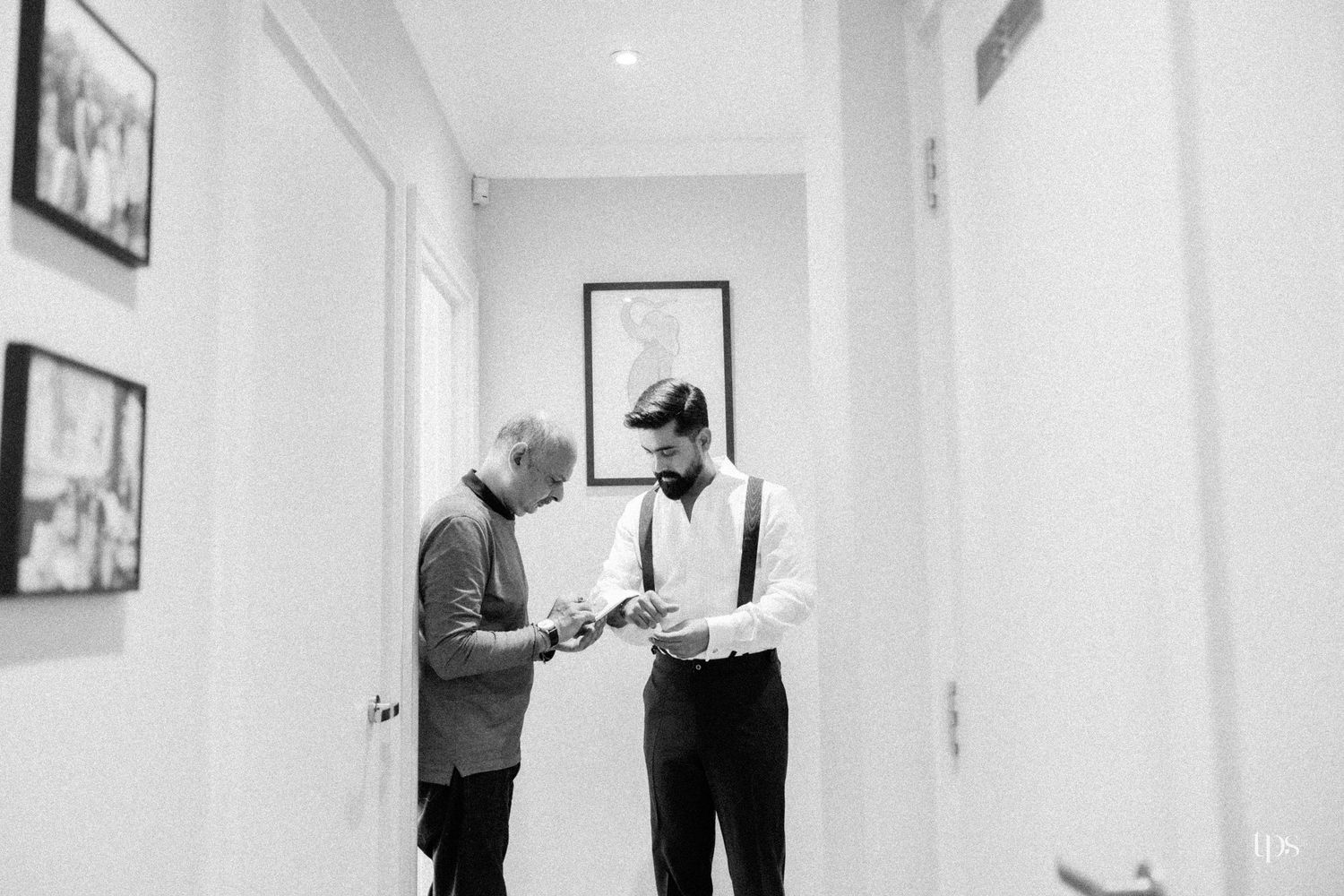 Black and white photo of groom and older man examining cufflinks in a hallway before a wedding ceremony.