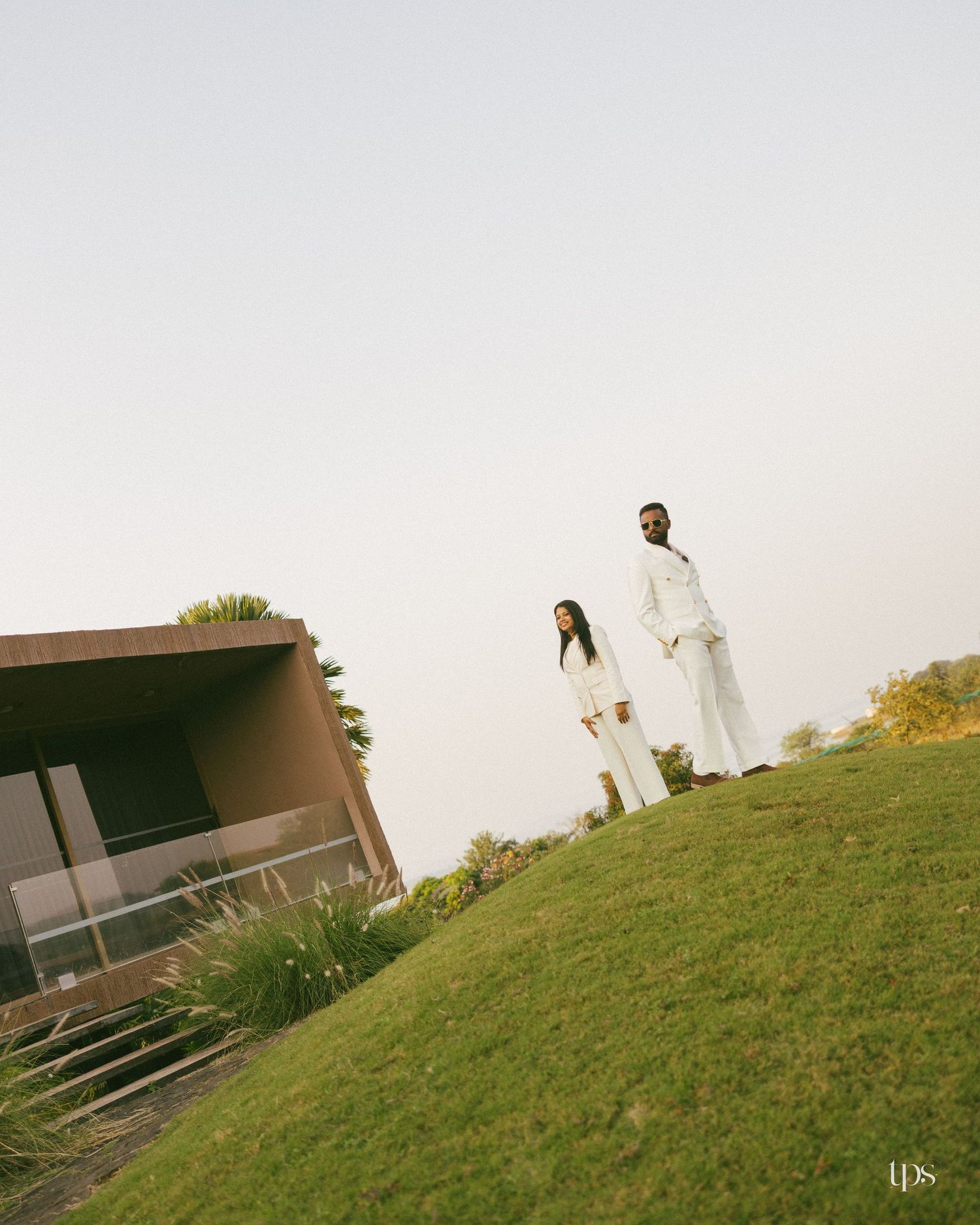 Two people in matching white outfits stand on a grassy hill beside a modern brown building under a soft, hazy sky.