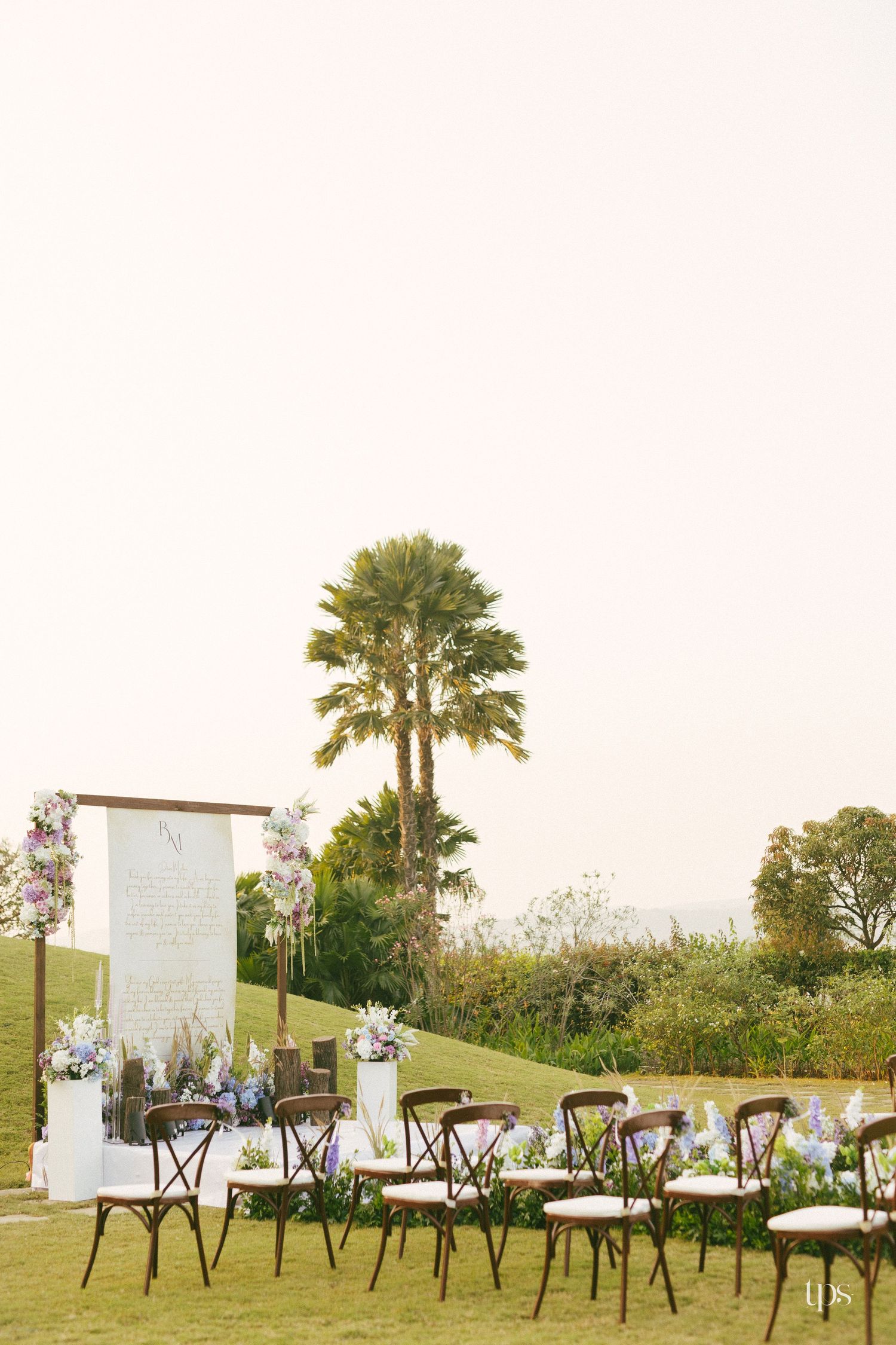 Outdoor garden wedding ceremony setup with wooden cross-back chairs, floral arrangements, and a welcome sign beneath a palm tree at sunset.
