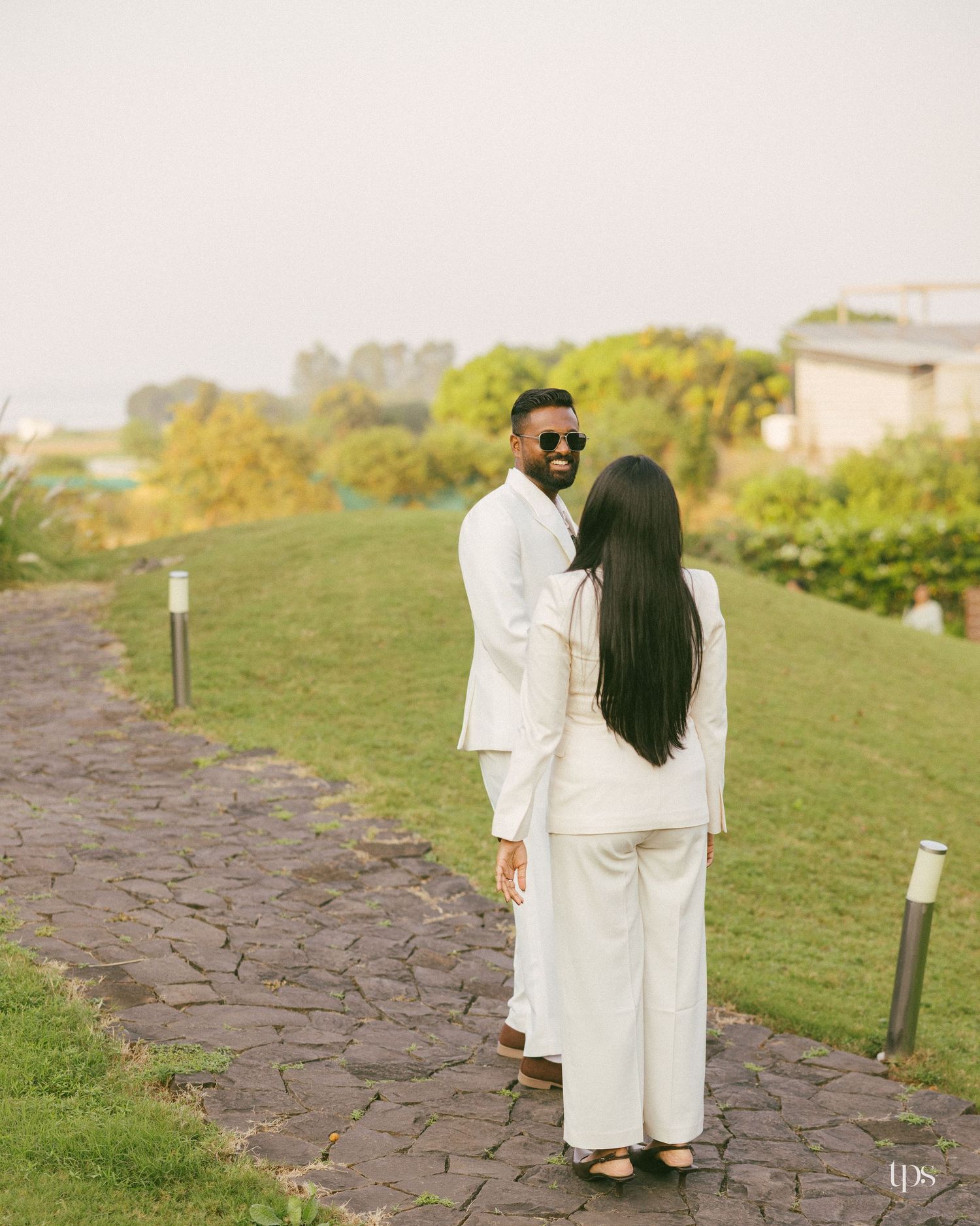 Couple in matching white outfits stand facing each other on a stone path beside a grassy hillside with trees and a building behind.