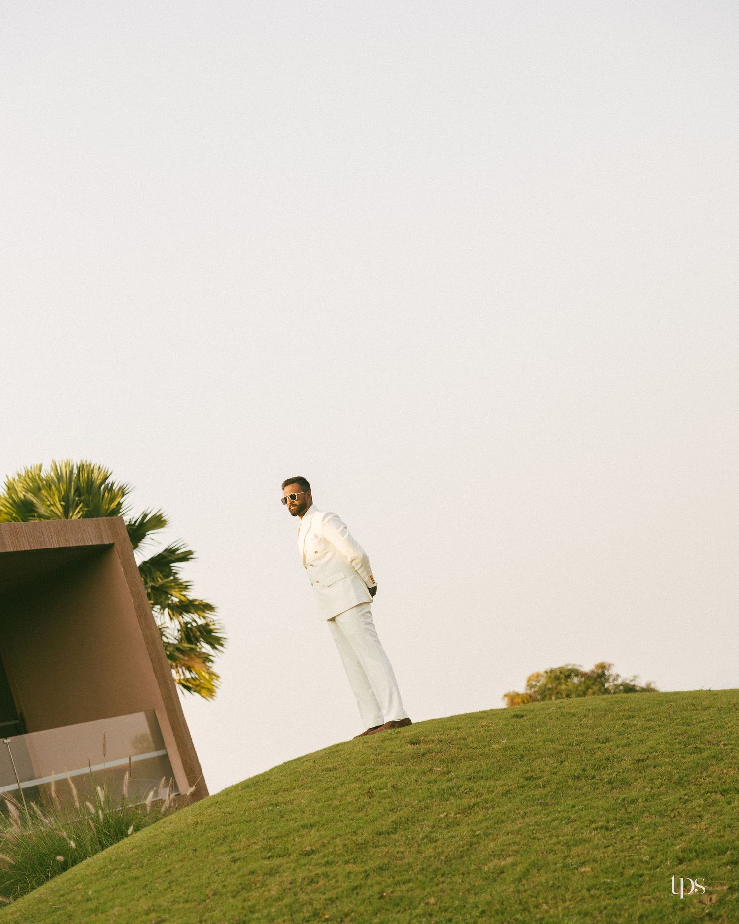 Man in a white suit and sunglasses stands on a grassy hill beside a modern brown building and palm tree under a pale sky.