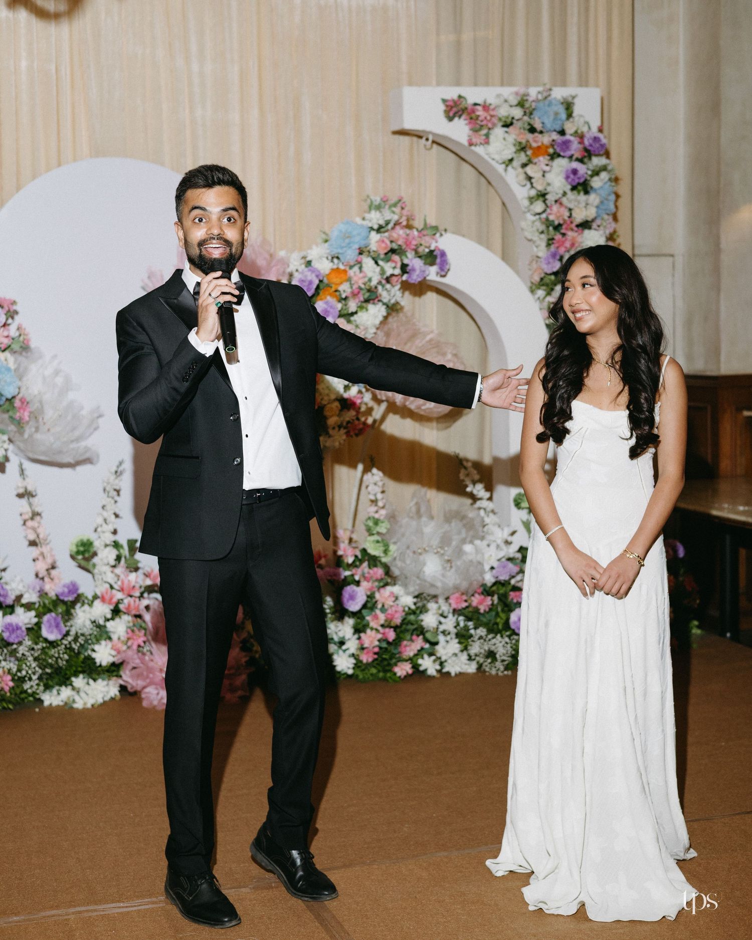 A man in a black suit speaks into a microphone beside a woman in a white gown at a wedding reception with pastel floral arches.
