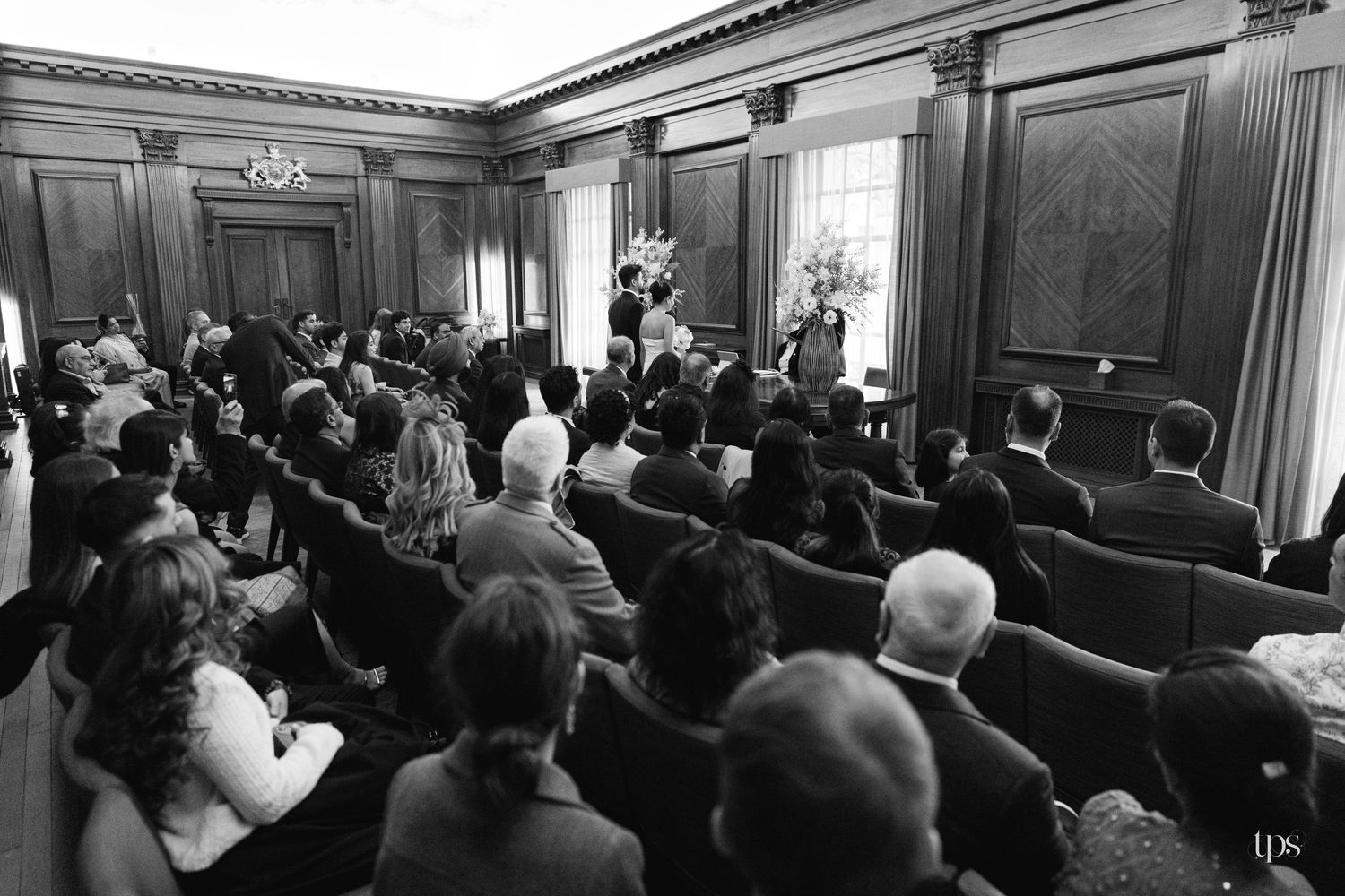 Black and white photo of wedding ceremony guests seated in elegant wood-paneled room watching couple at altar.