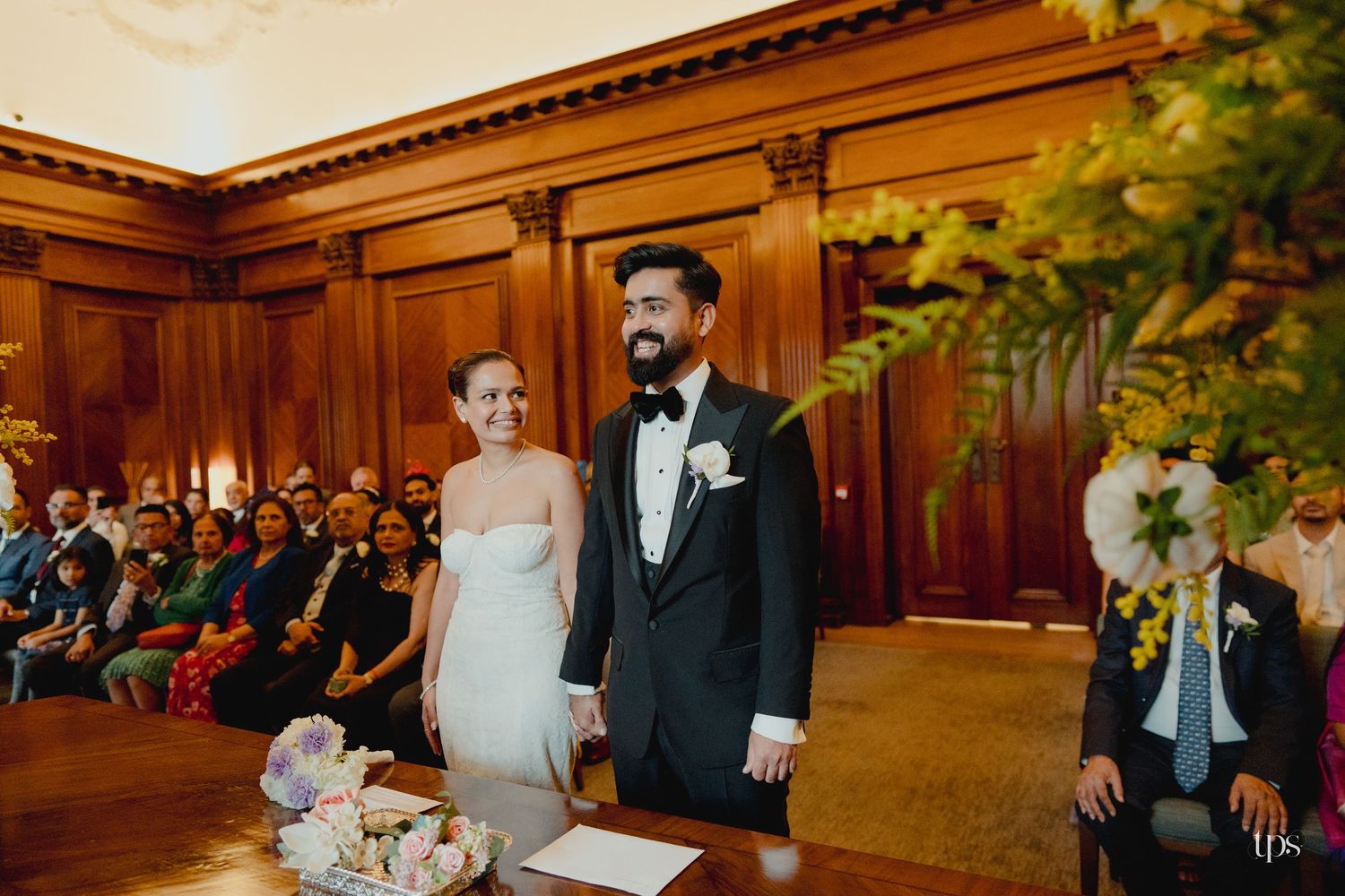 Bride and groom holding hands at wedding ceremony altar in elegant wood-paneled room with guests seated behind them.