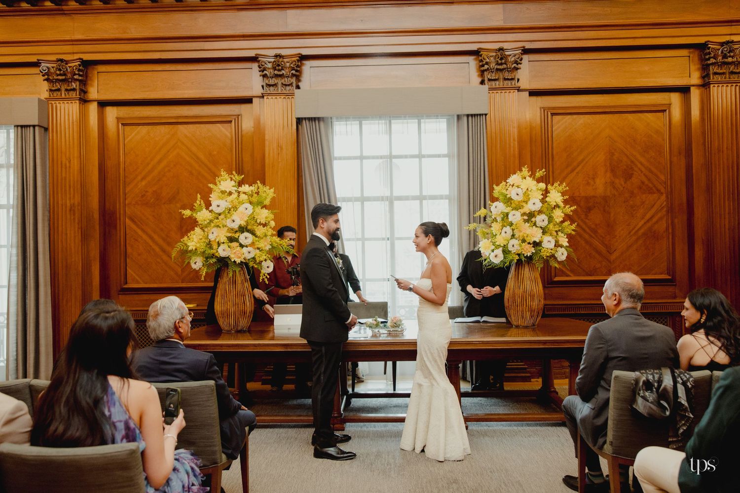 Couple exchanging vows at elegant indoor wedding ceremony with wood paneling and yellow floral arrangements.