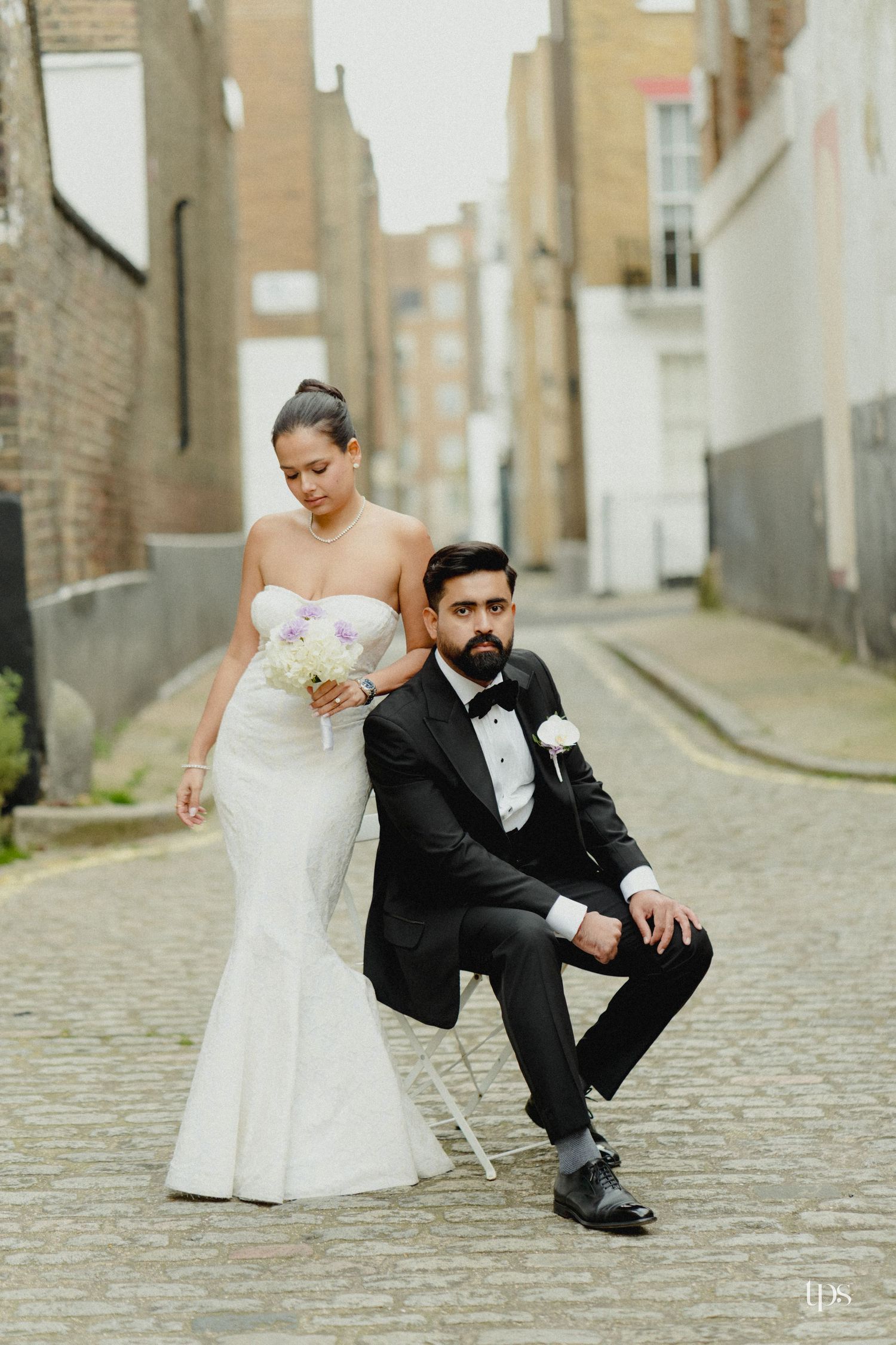 Bride in white mermaid gown and groom in black tuxedo pose on cobblestone alley for wedding portrait.
