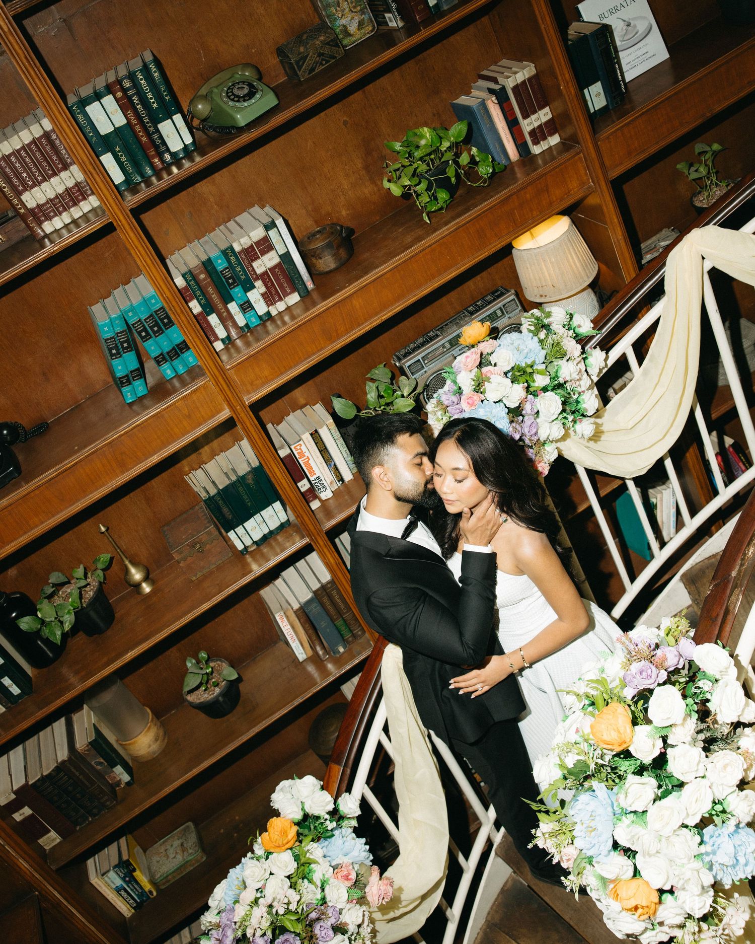 Bride and groom embrace on a library staircase surrounded by bookshelves and lush pastel wedding floral arrangements.