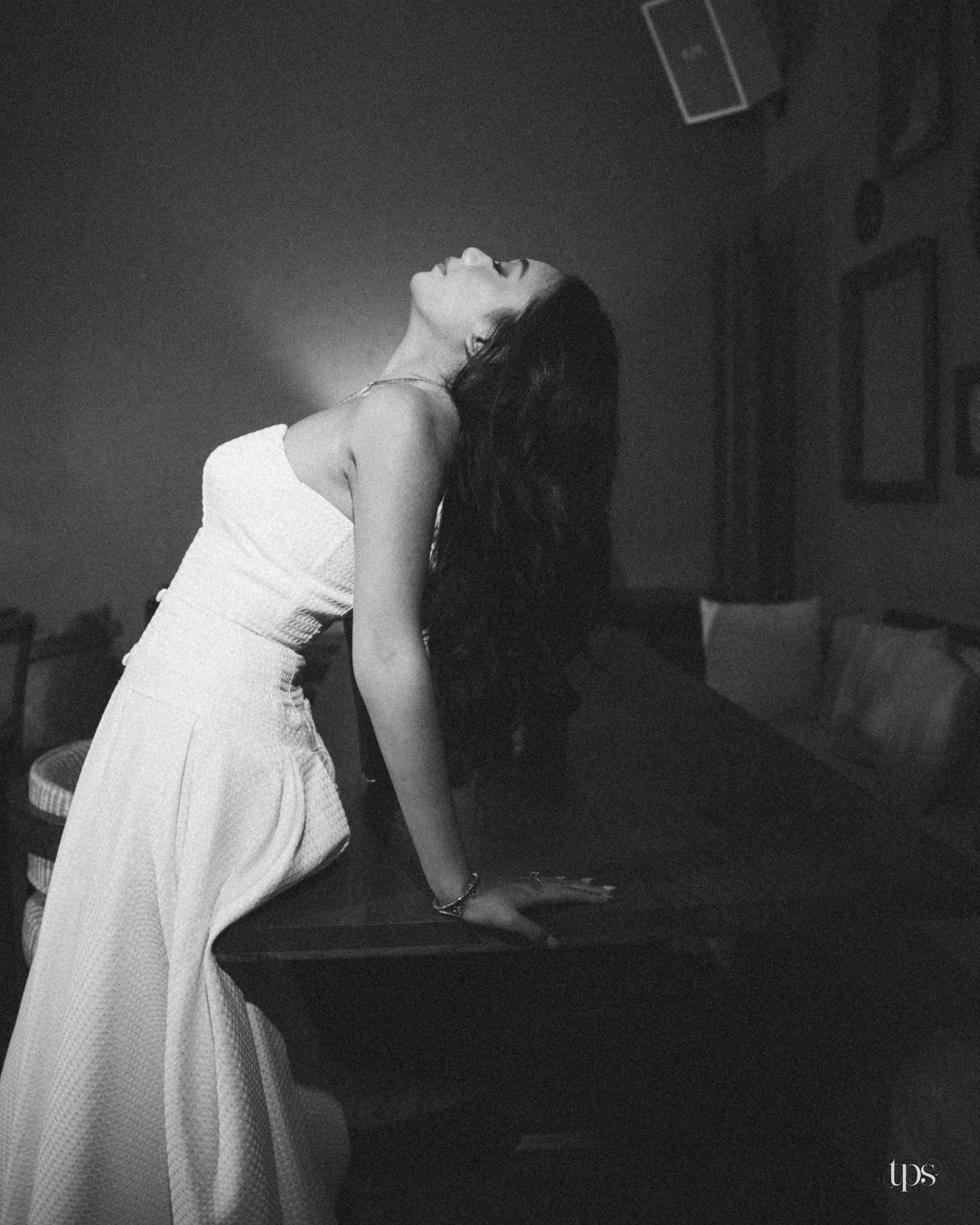 Black and white portrait of a woman in a white strapless dress leaning back against a wooden table with her head tilted up in a dim room.