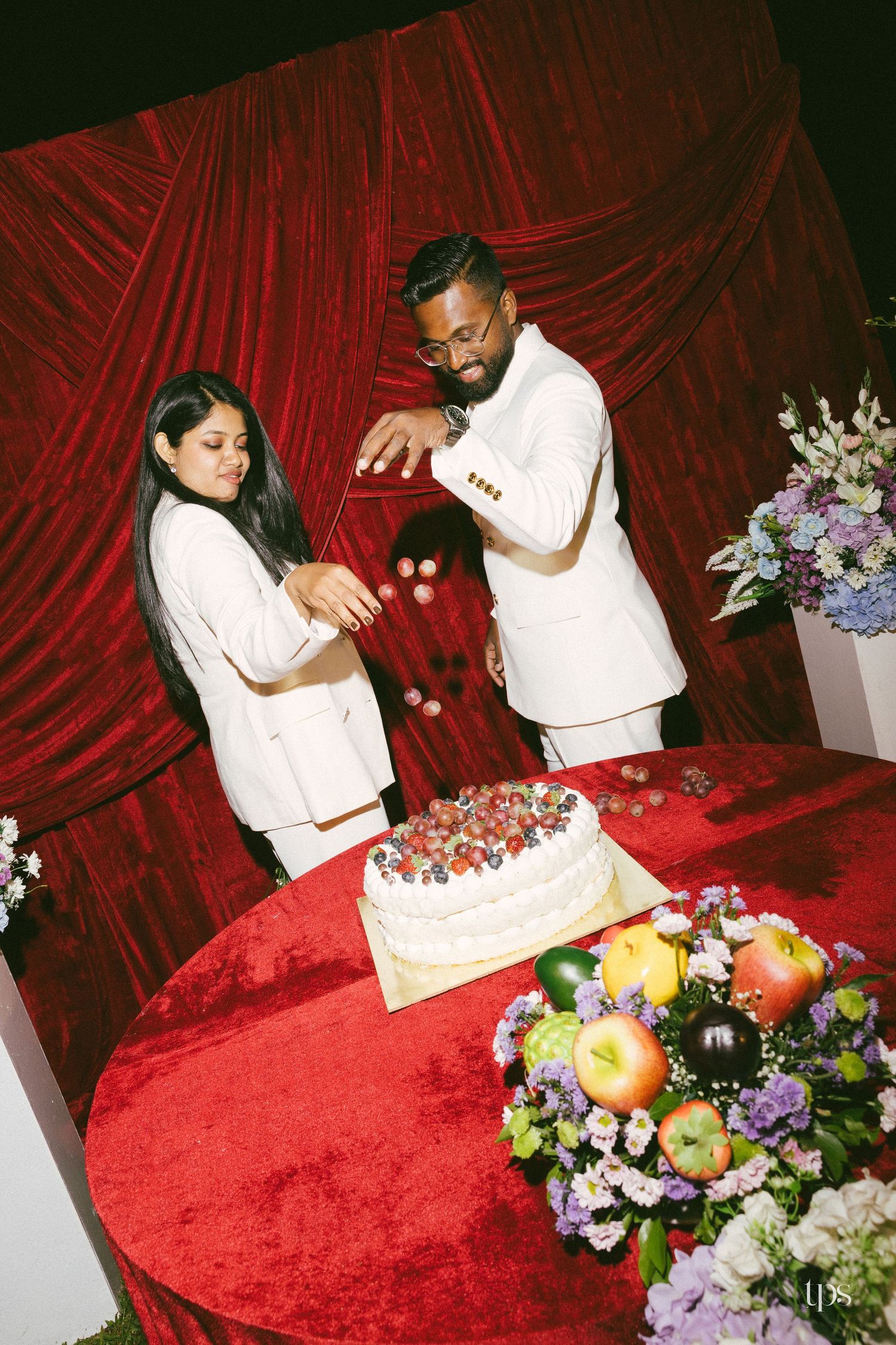 Two people in white outfits toss berries over a berry-topped white cake on a red velvet table with fruit and flowers.