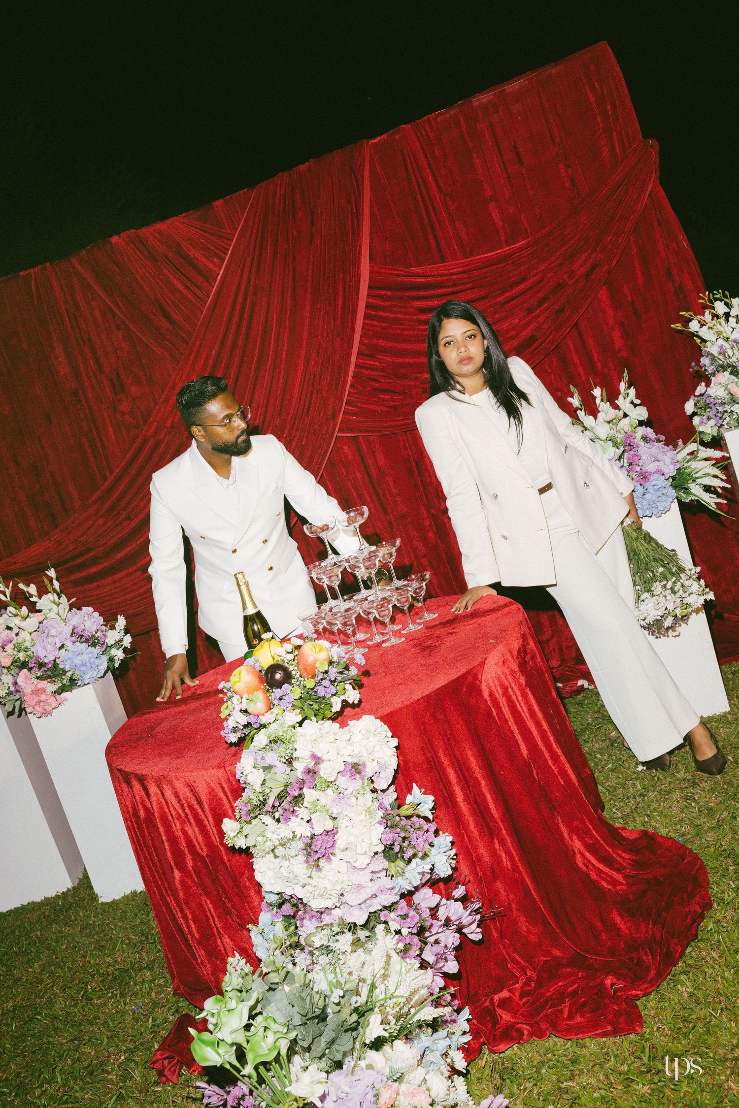 Couple in white outfits stand beside a red velvet table with a champagne tower and pastel floral arrangements before a red draped backdrop.
