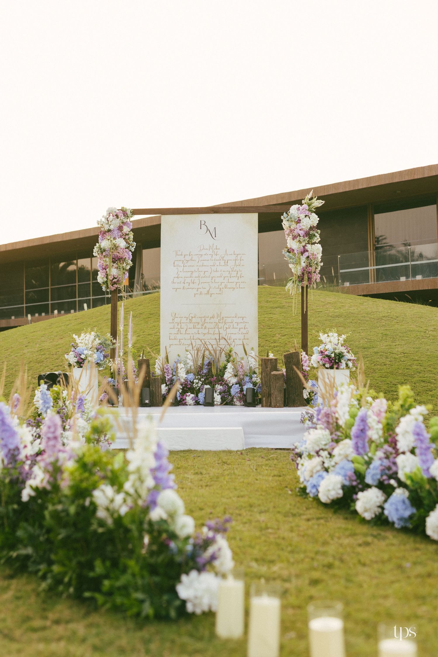 Outdoor wedding ceremony setup on a grassy hillside with a sign, floral arches, blue and white blooms, and candles beside a modern building.
