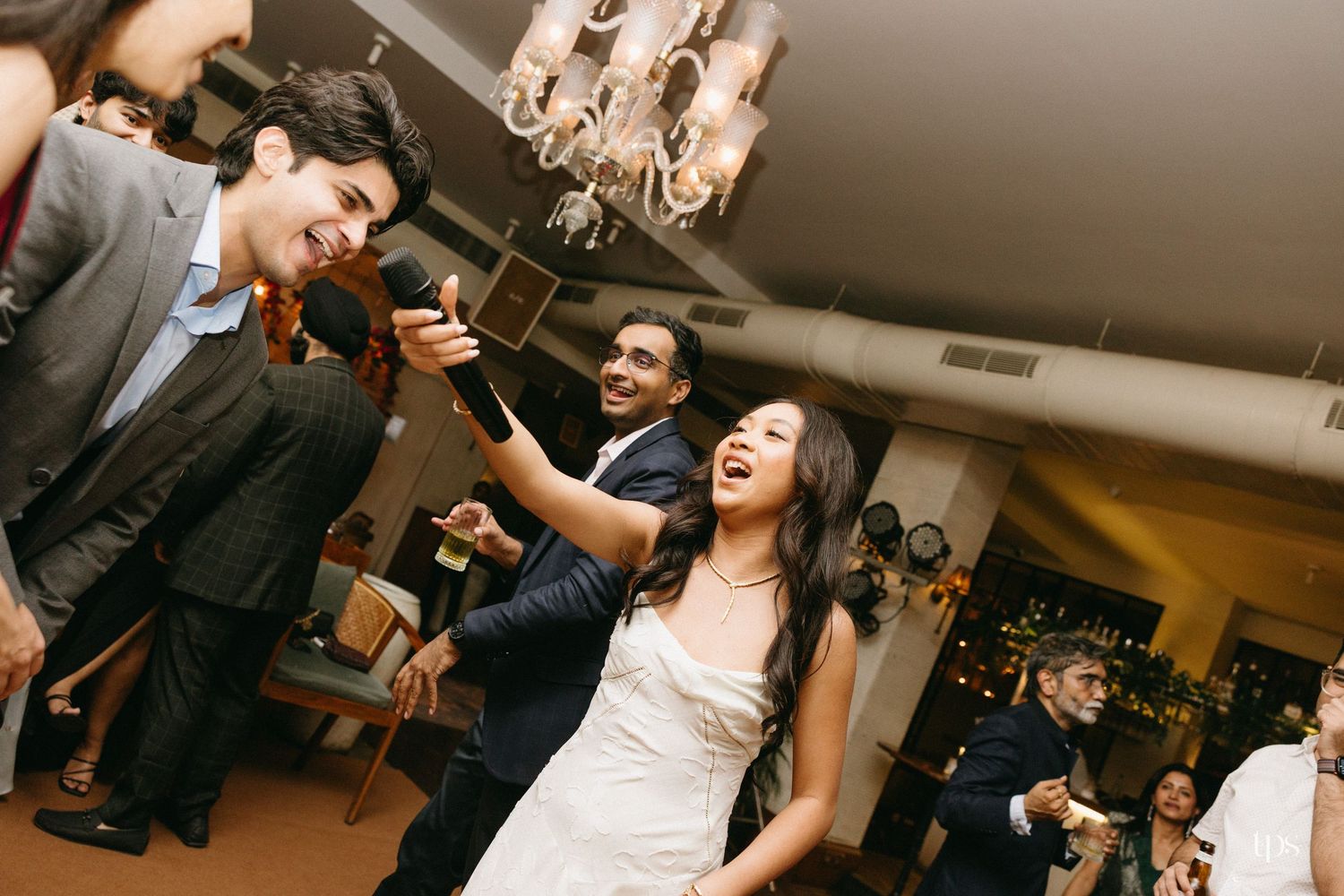 Bride in a white gown sings into a microphone while smiling guests dance and celebrate around her at a lively wedding reception.