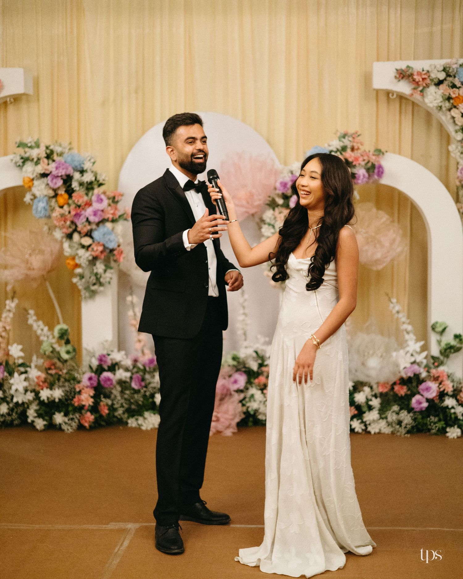 Bride in a white gown and groom in a black tuxedo smile while sharing a microphone in front of floral arches at a wedding reception.