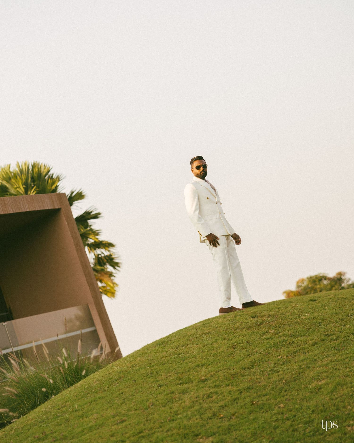 Man in a white double-breasted suit and sunglasses stands on a grassy hill beside a modern building and palm tree against a hazy sky.