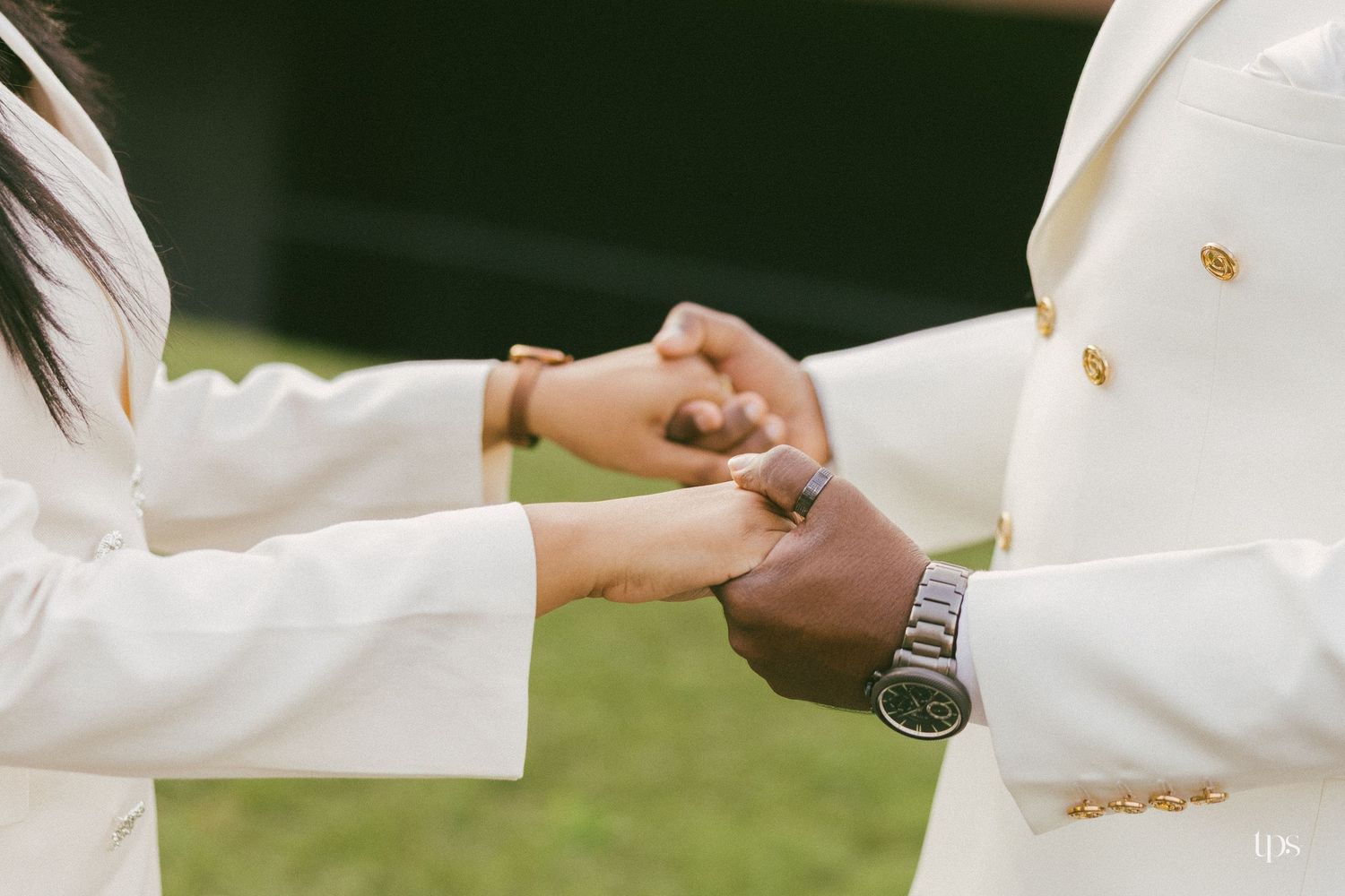 Two people in matching cream blazers with gold buttons hold hands outdoors on a grassy lawn, one wearing a silver wristwatch.