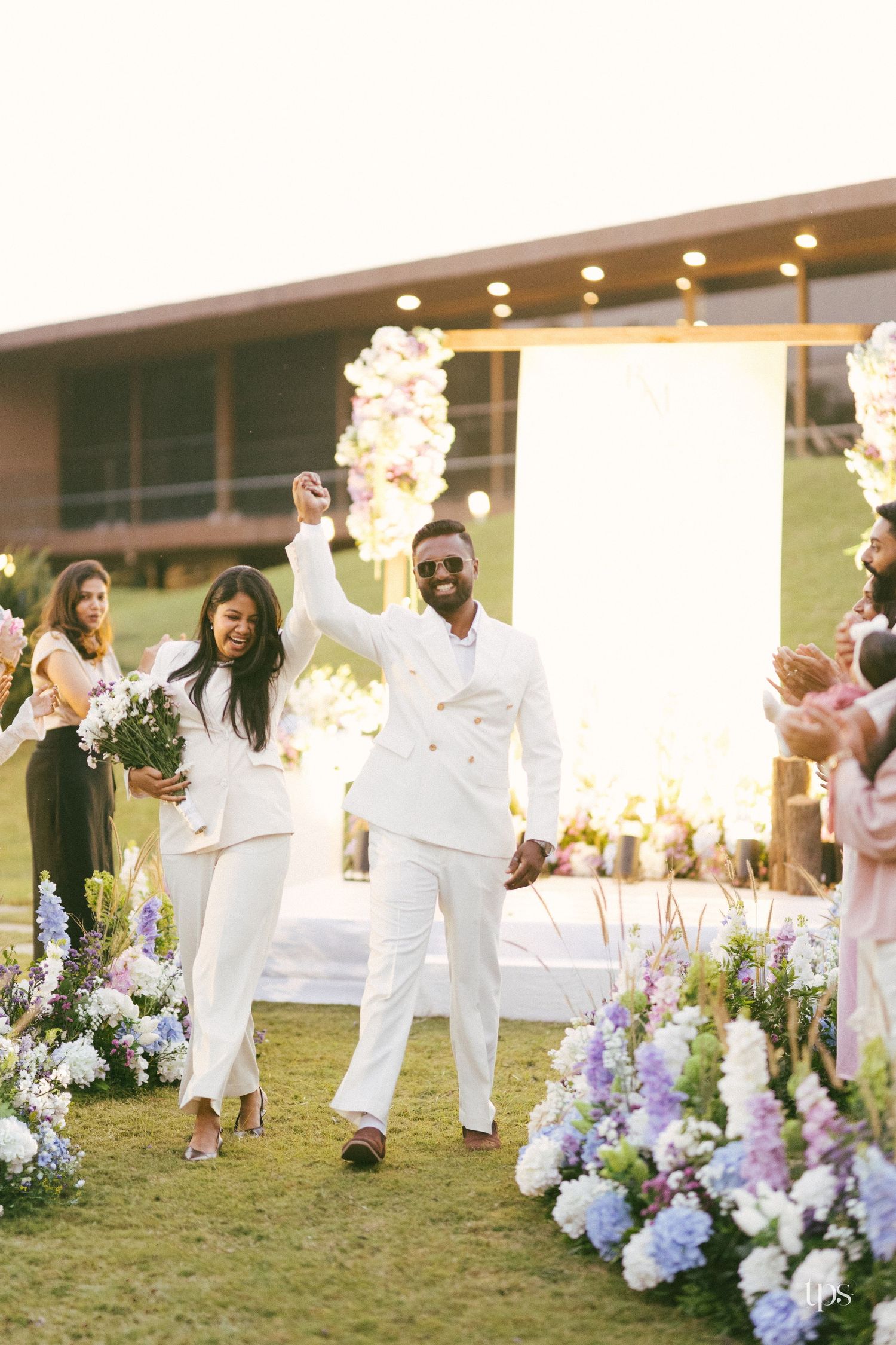 Bride and groom in matching white suits walk joyfully down a grassy aisle lined with pastel flowers at an outdoor wedding ceremony.