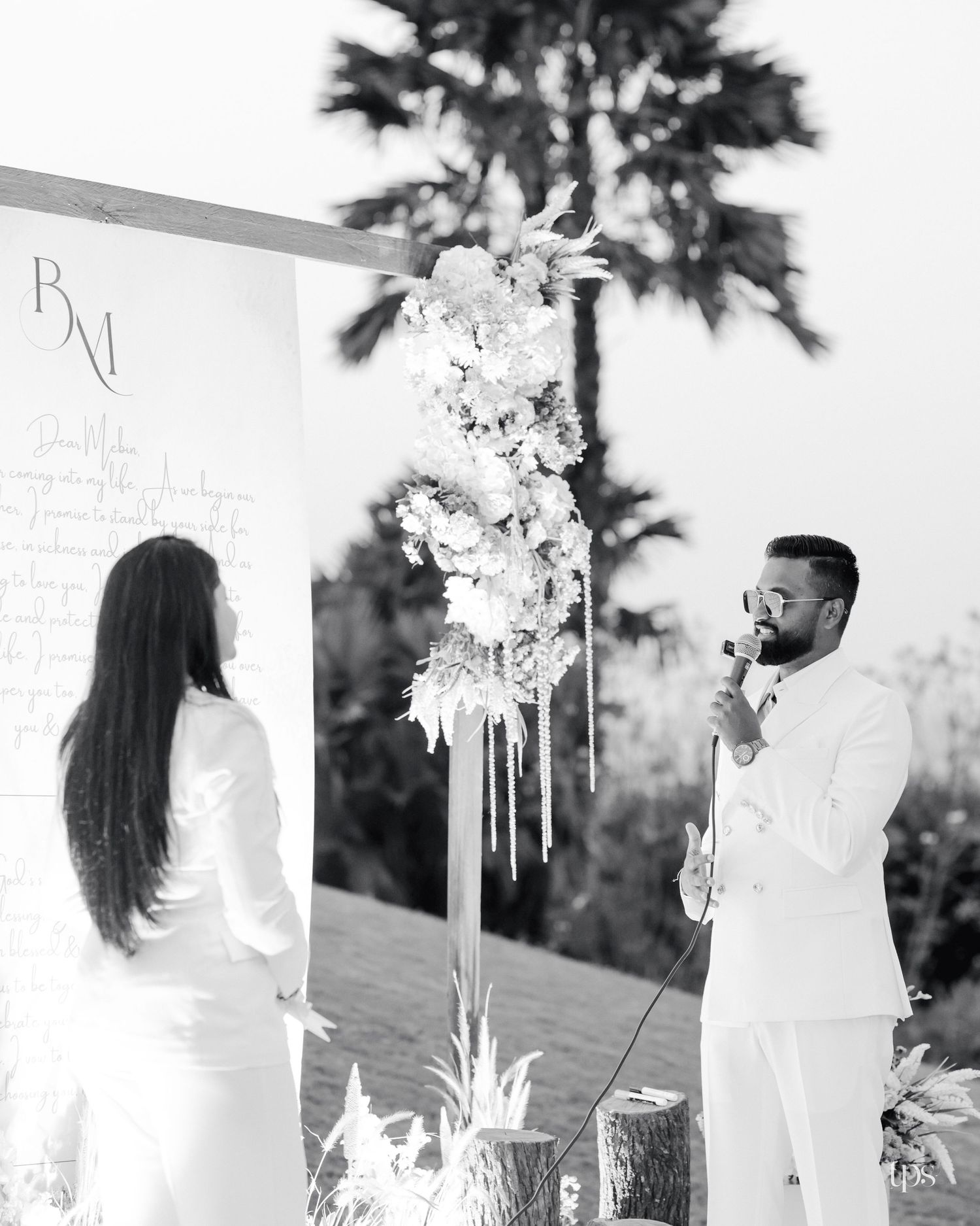 Black and white photo of a groom in a light suit speaking into a microphone beside a bride at an outdoor ceremony with white floral decor.
