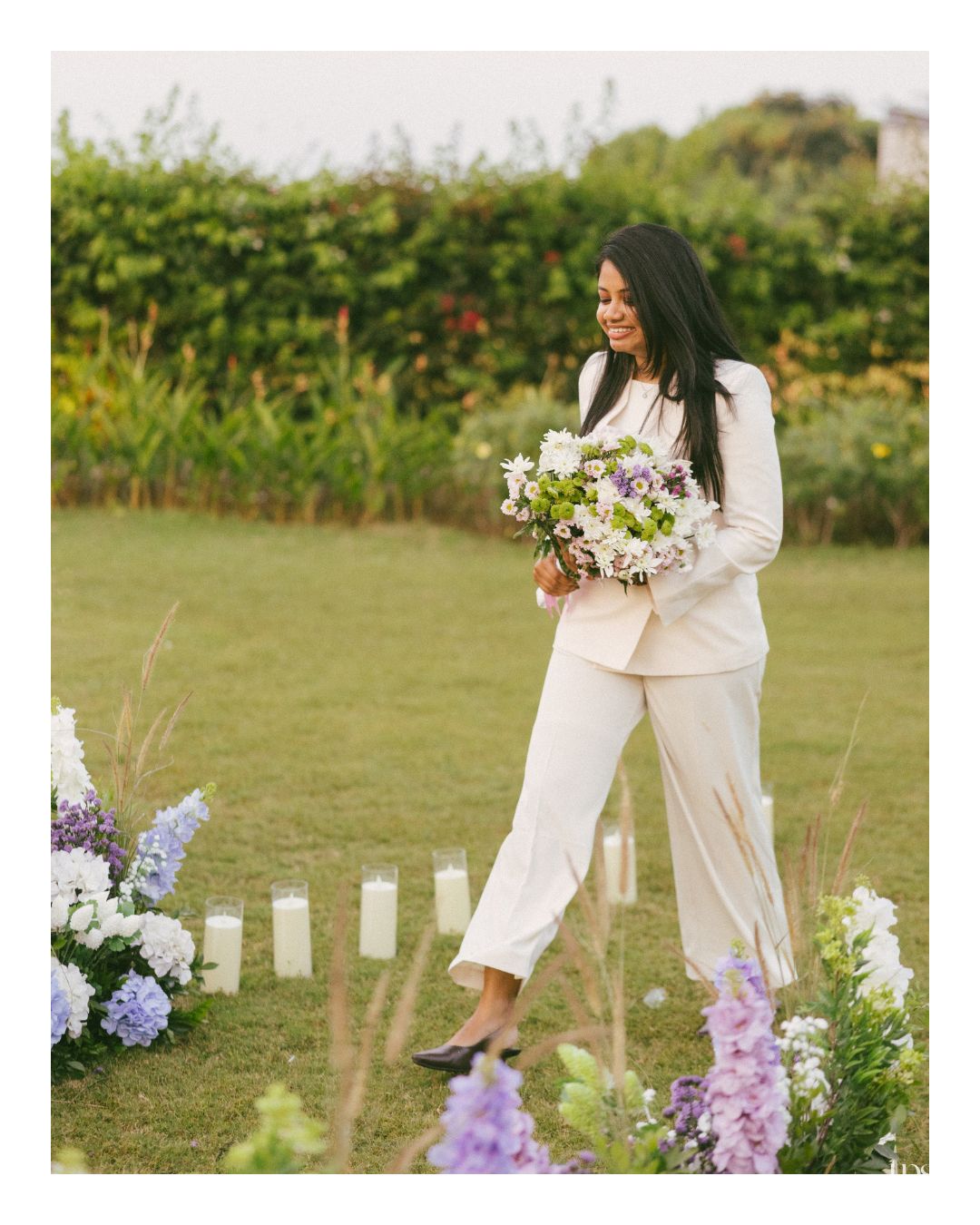 Smiling bride in a white pantsuit carrying a purple and white floral bouquet across a green lawn lined with candles and wildflowers.