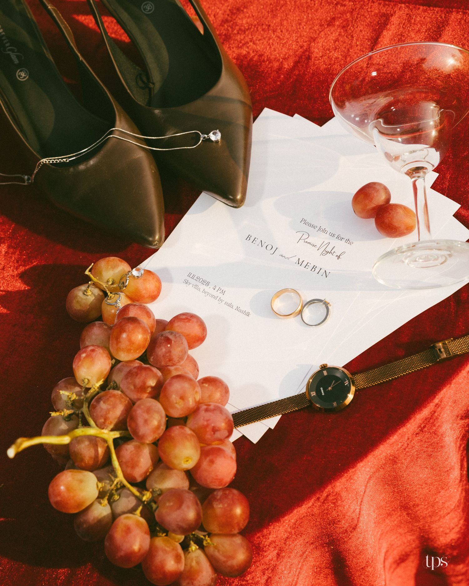 Wedding invitation, two gold rings, olive heels, a wristwatch, red grapes and a coupe glass styled on a red cloth.