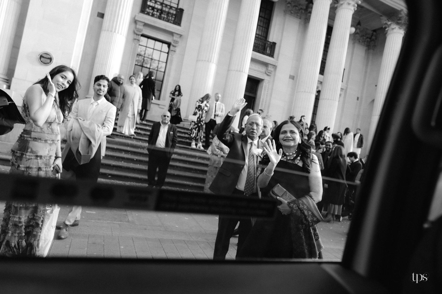 Black and white photo of wedding guests waving and celebrating outside a grand building with tall columns.