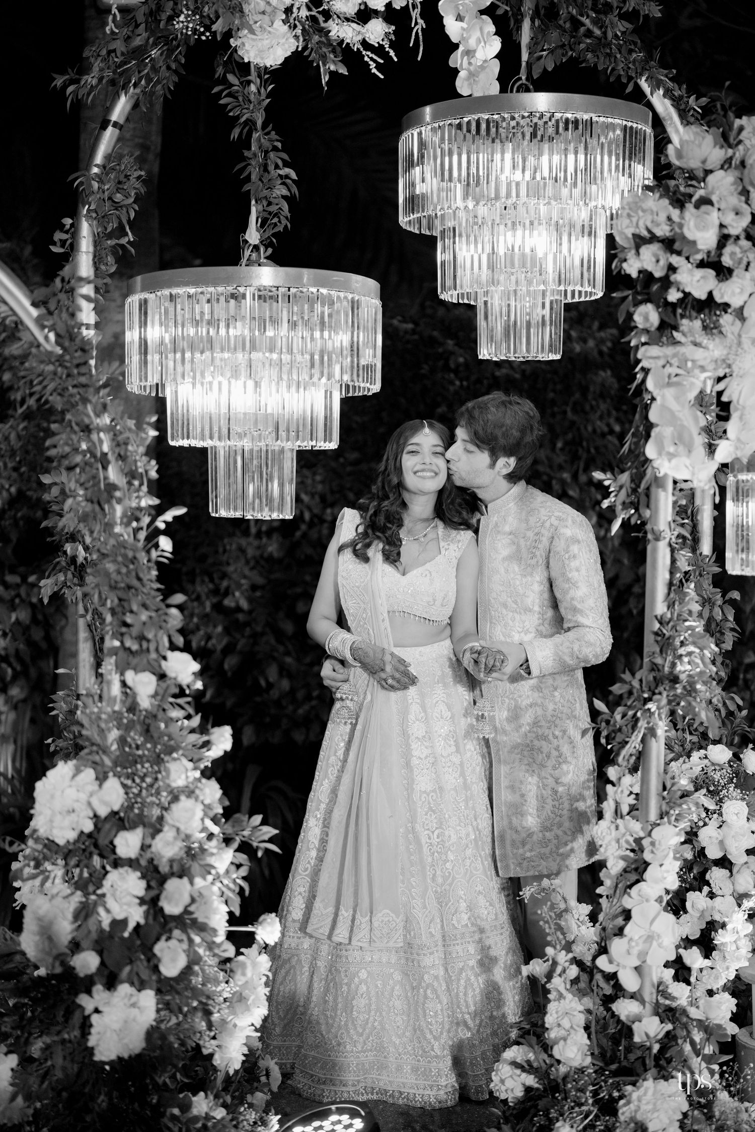 Black and white photo of a couple in traditional Indian attire posing under crystal chandeliers surrounded by floral arrangements.