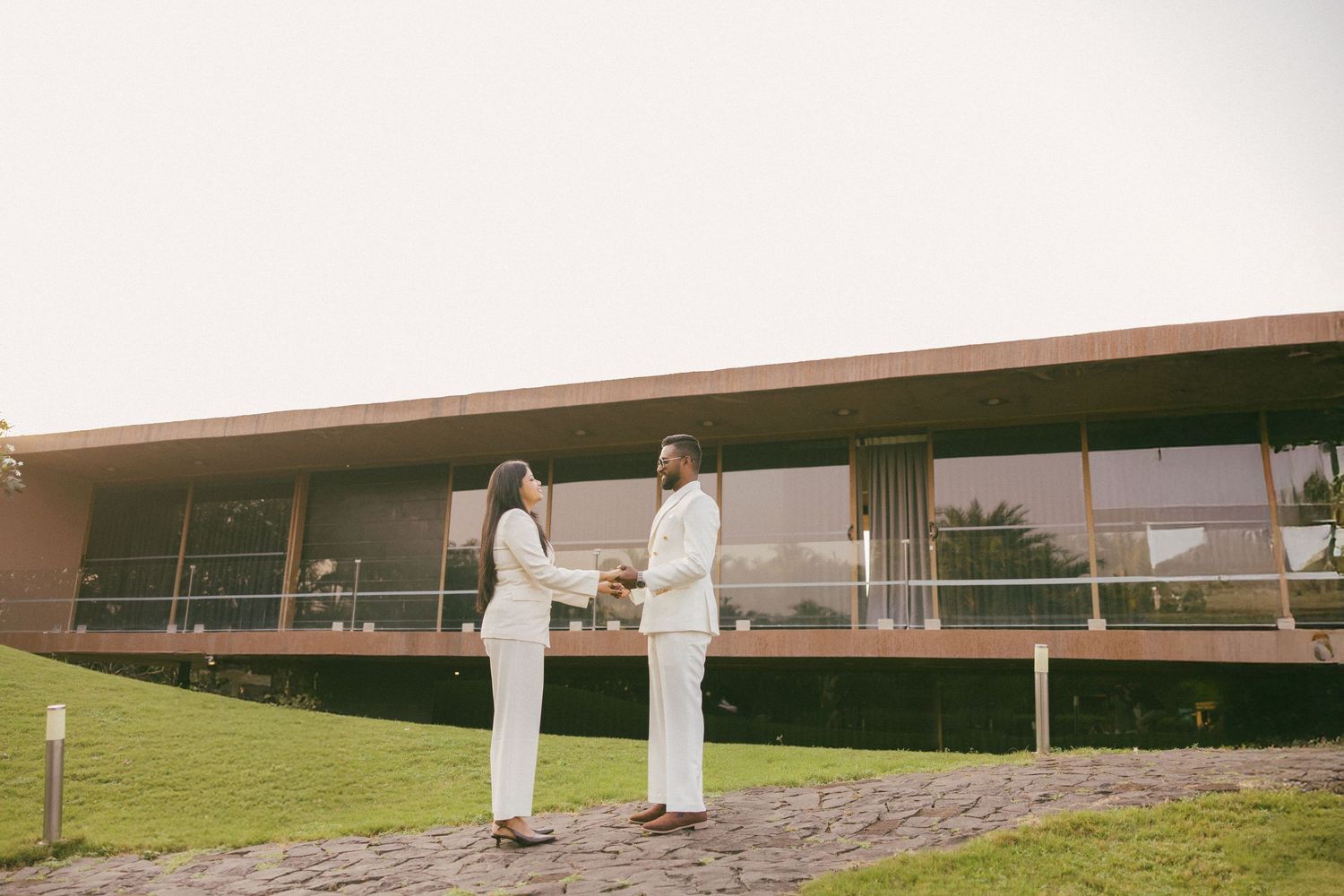 Couple in matching white outfits holding hands on a stone path in front of a modern glass-walled building surrounded by green lawn.