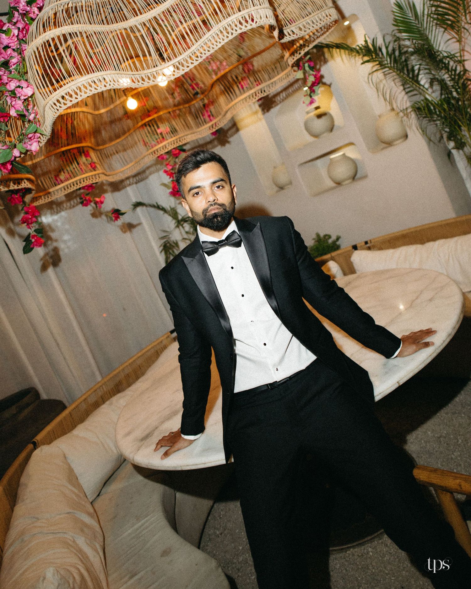 Man in a black tuxedo and bow tie leans casually against a round marble table beneath a woven rattan chandelier with pink flowers.
