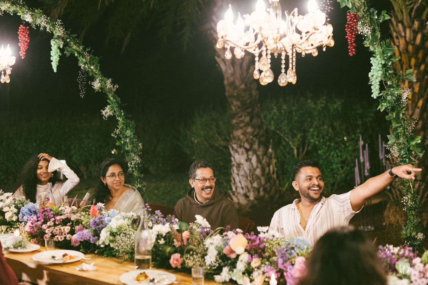 Smiling guests laugh and point while seated at a long outdoor dinner table with flower arrangements under a sparkling chandelier at night.