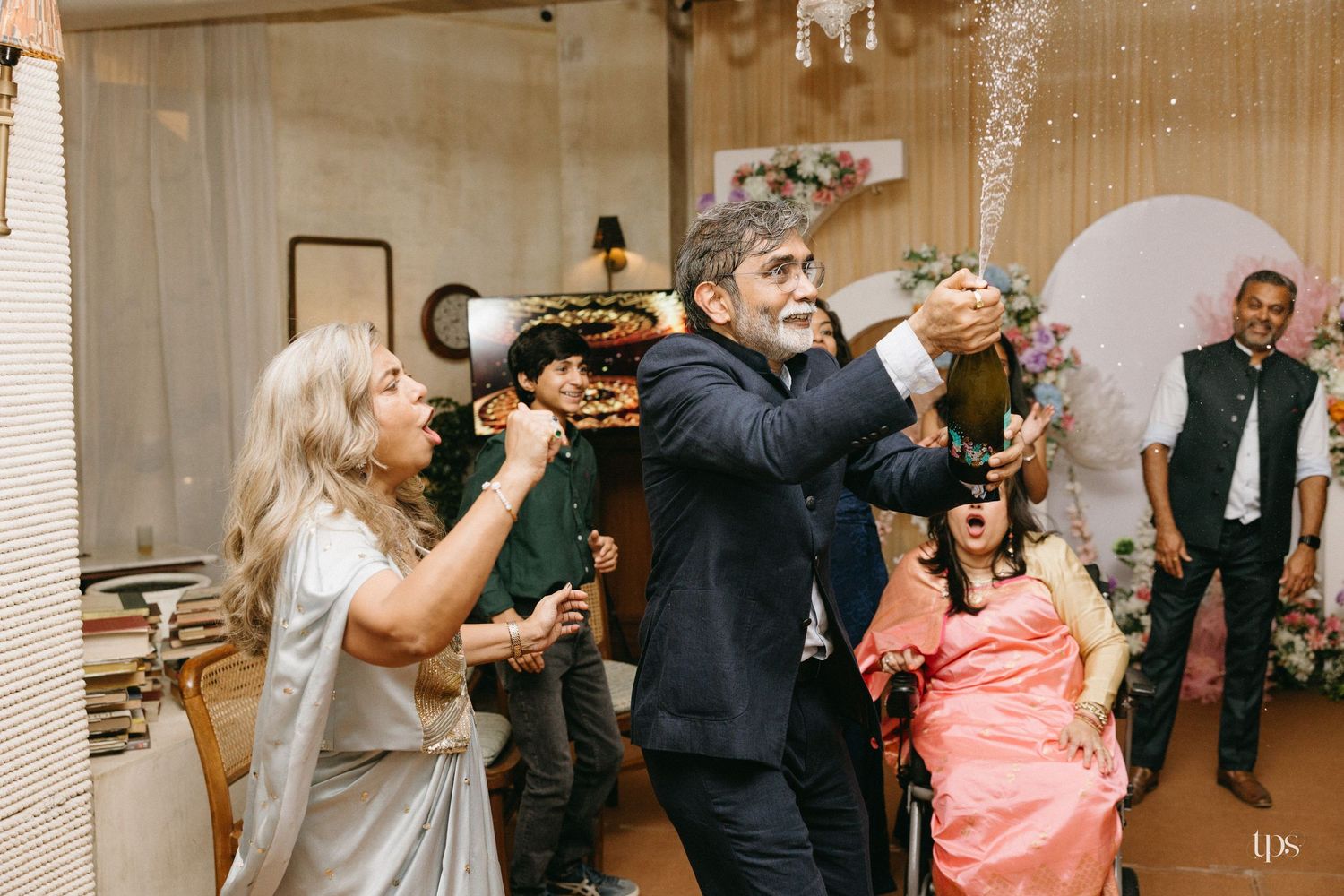 Guests celebrate at an indoor wedding reception as a man in a dark suit pops a champagne bottle beside a woman in a silver gown.