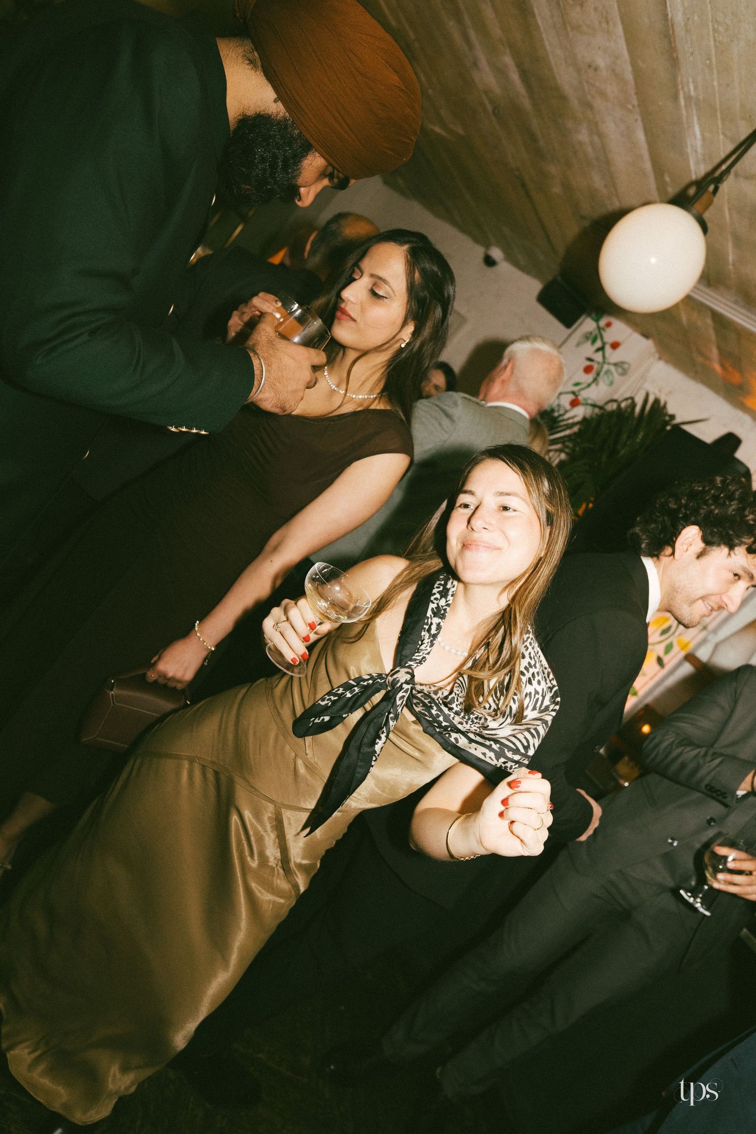 People socializing at an indoor party, with women in elegant attire holding drinks under warm ambient lighting.