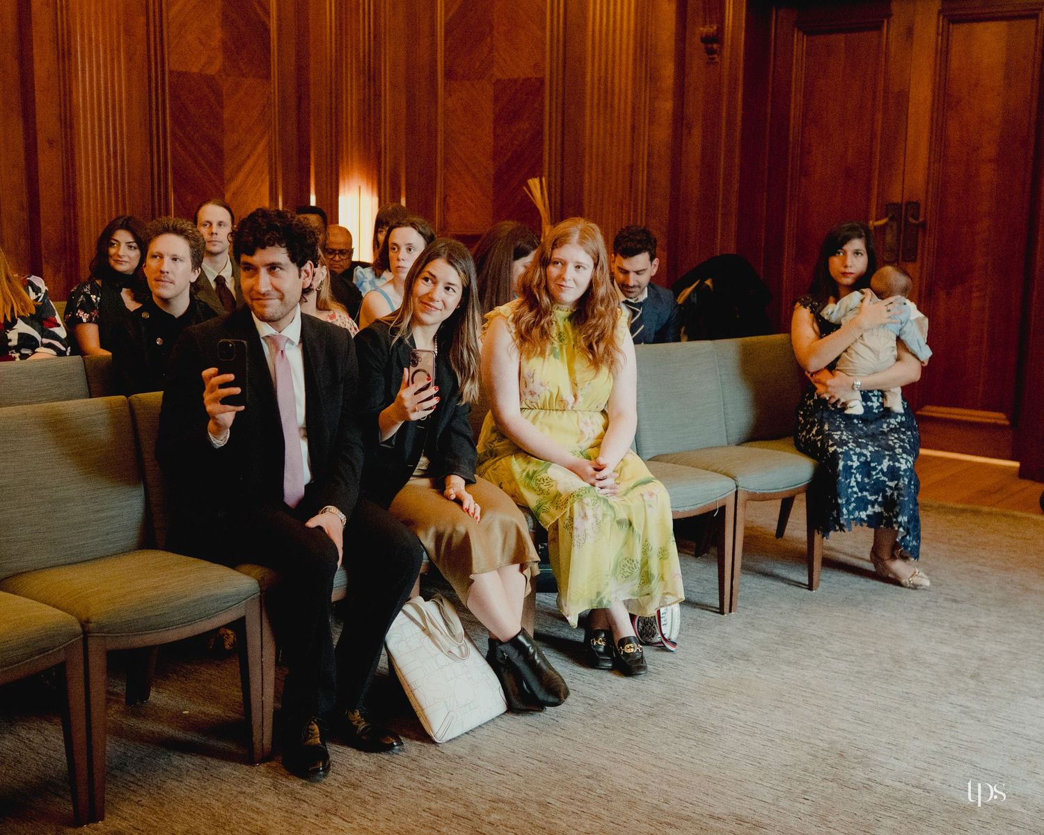 Guests seated in wooden pews at an elegant indoor ceremony, smiling and dressed in formal attire.